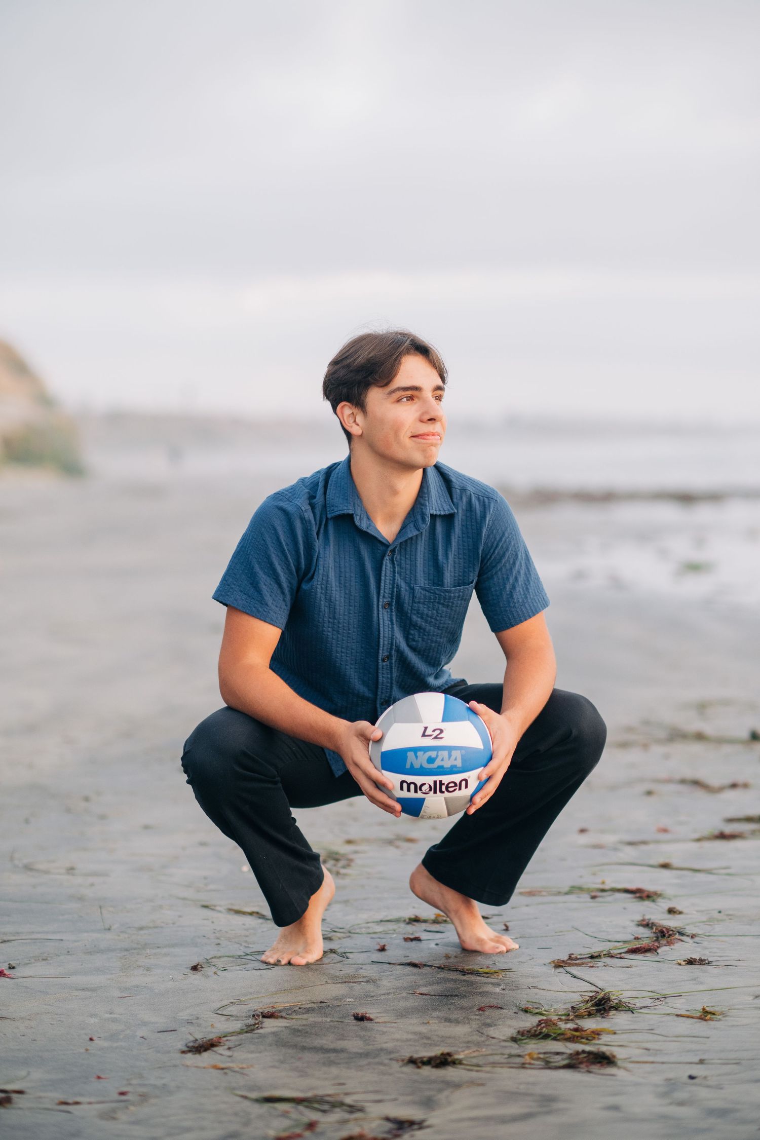 Young man crouching on beach holding Molten NCAA volleyball, wearing blue shirt and black pants.