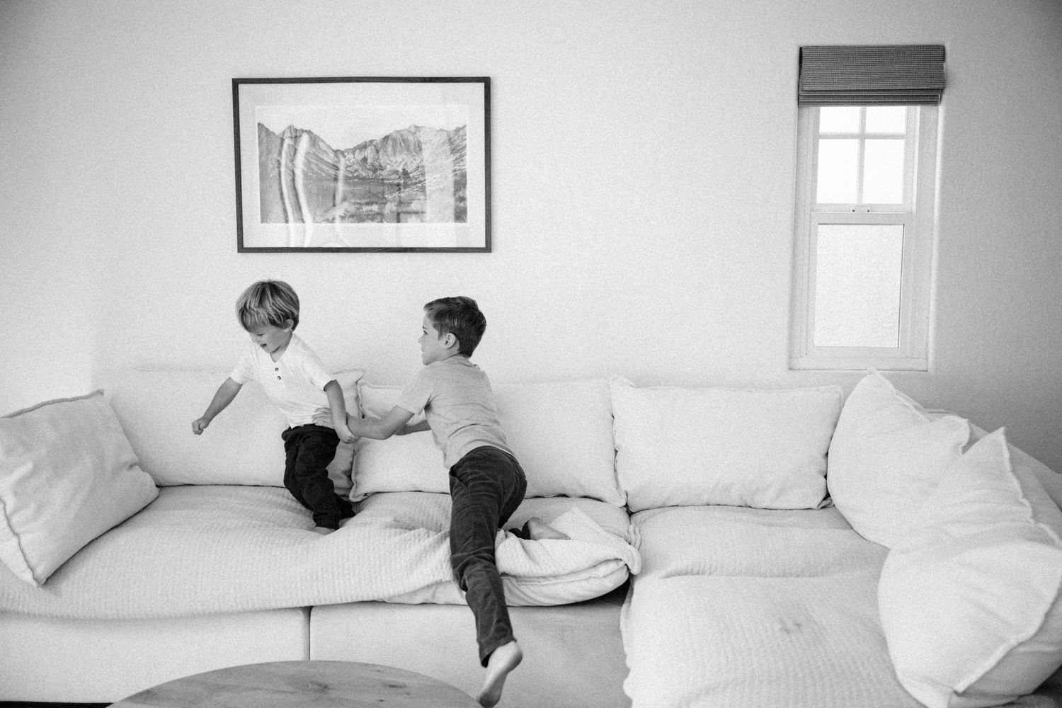Two children playing on a large white sectional sofa in a bright living room, black and white photo.