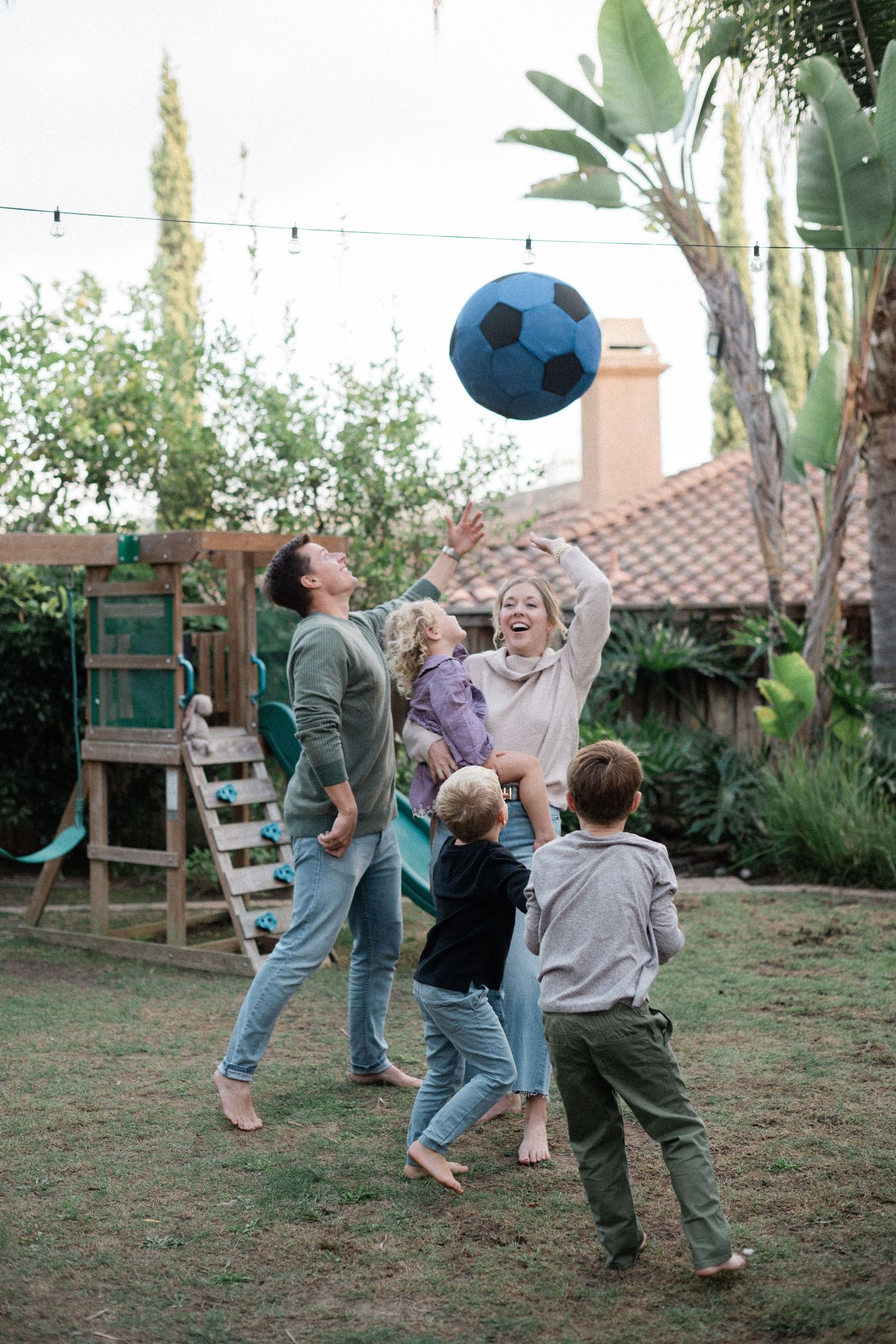Happy family of five playing with a large soccer ball together in a lush backyard playground.