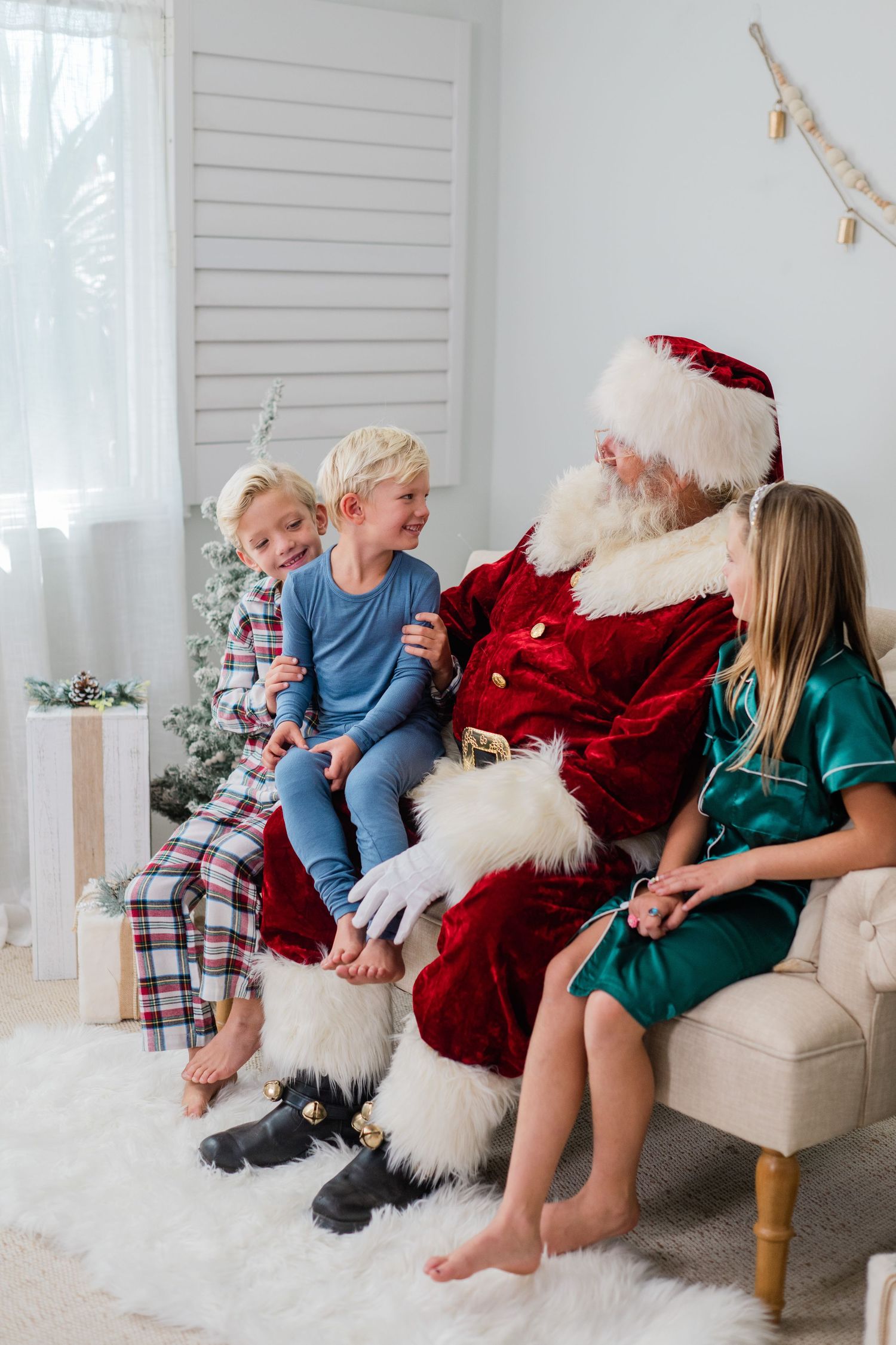 Santa Claus sits in a chair with three children in pajamas during a festive Christmas photo session in a bright white room.