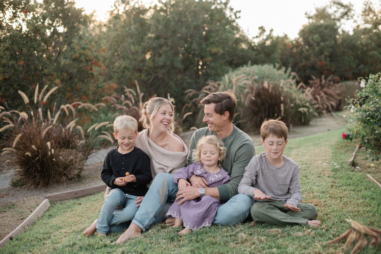 Happy family of five sitting together on green grass surrounded by lush garden greenery during golden hour.