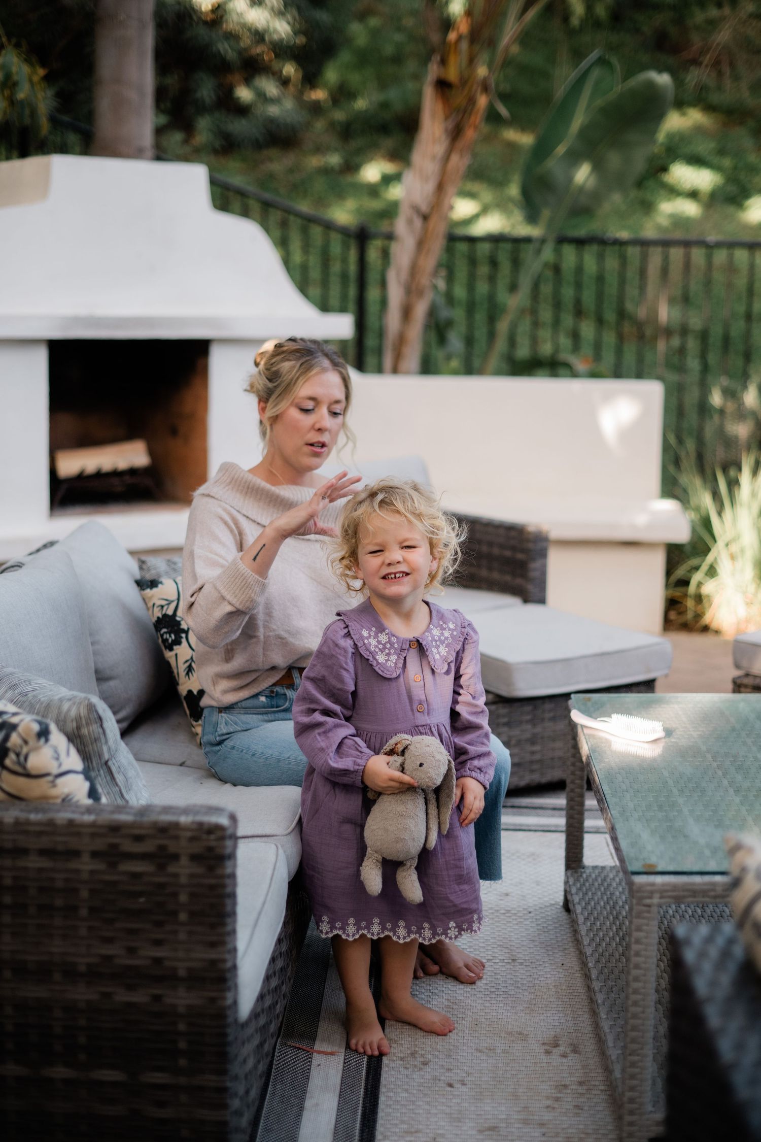 Mother styling toddler girl's hair on outdoor patio with fireplace, child holding stuffed bunny toy.