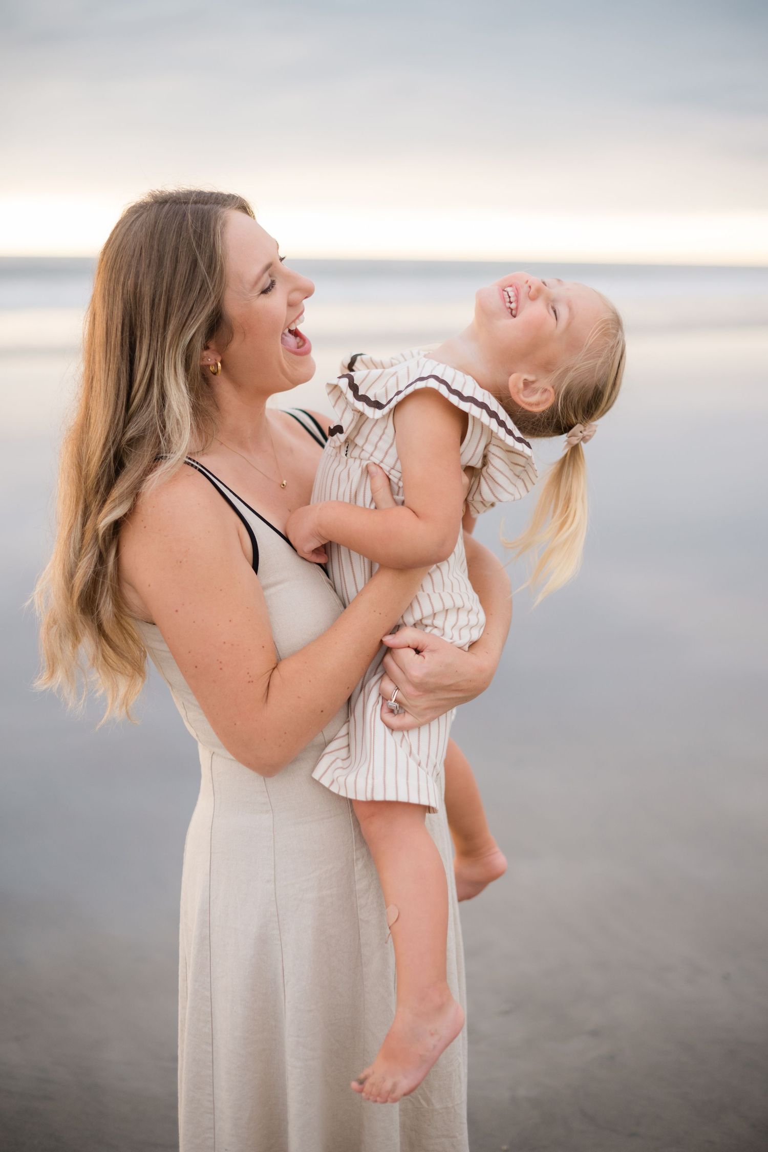 A laughing mother holds her giggling blonde toddler girl on a serene beach at sunset.