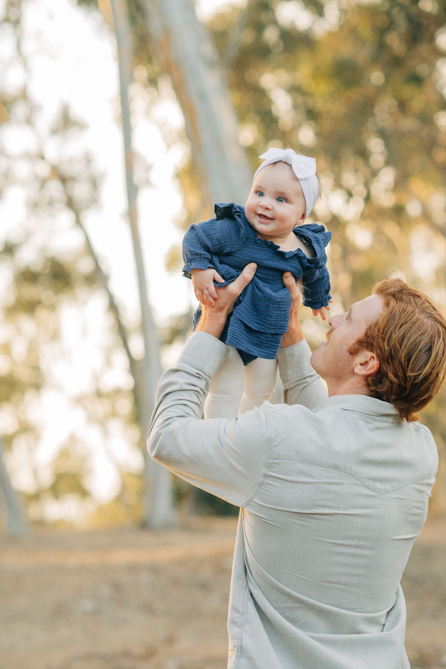 A joyful father lifts his smiling baby girl wearing a blue dress and white bow outdoors among trees.