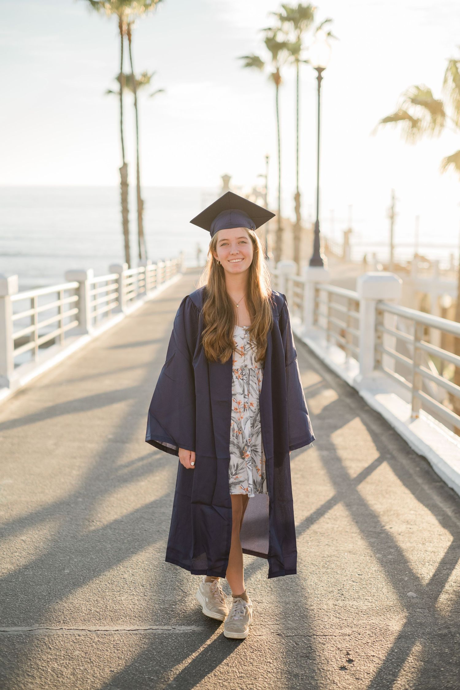 Smiling graduate in navy cap and gown poses on sunny boardwalk with palm trees and ocean backdrop.