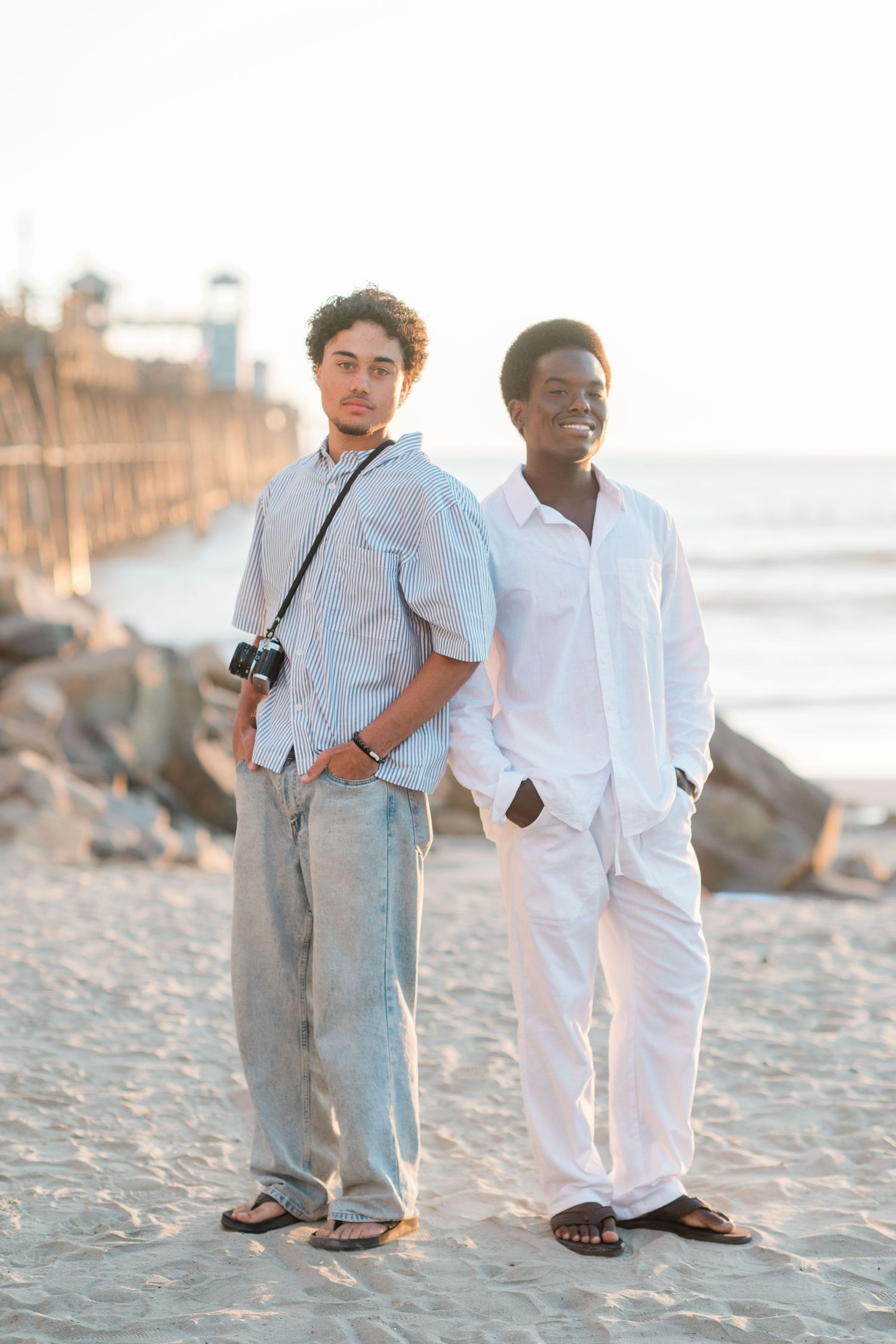 Two young men standing on a sandy beach near a pier, one wearing white and one in a striped shirt with a camera.