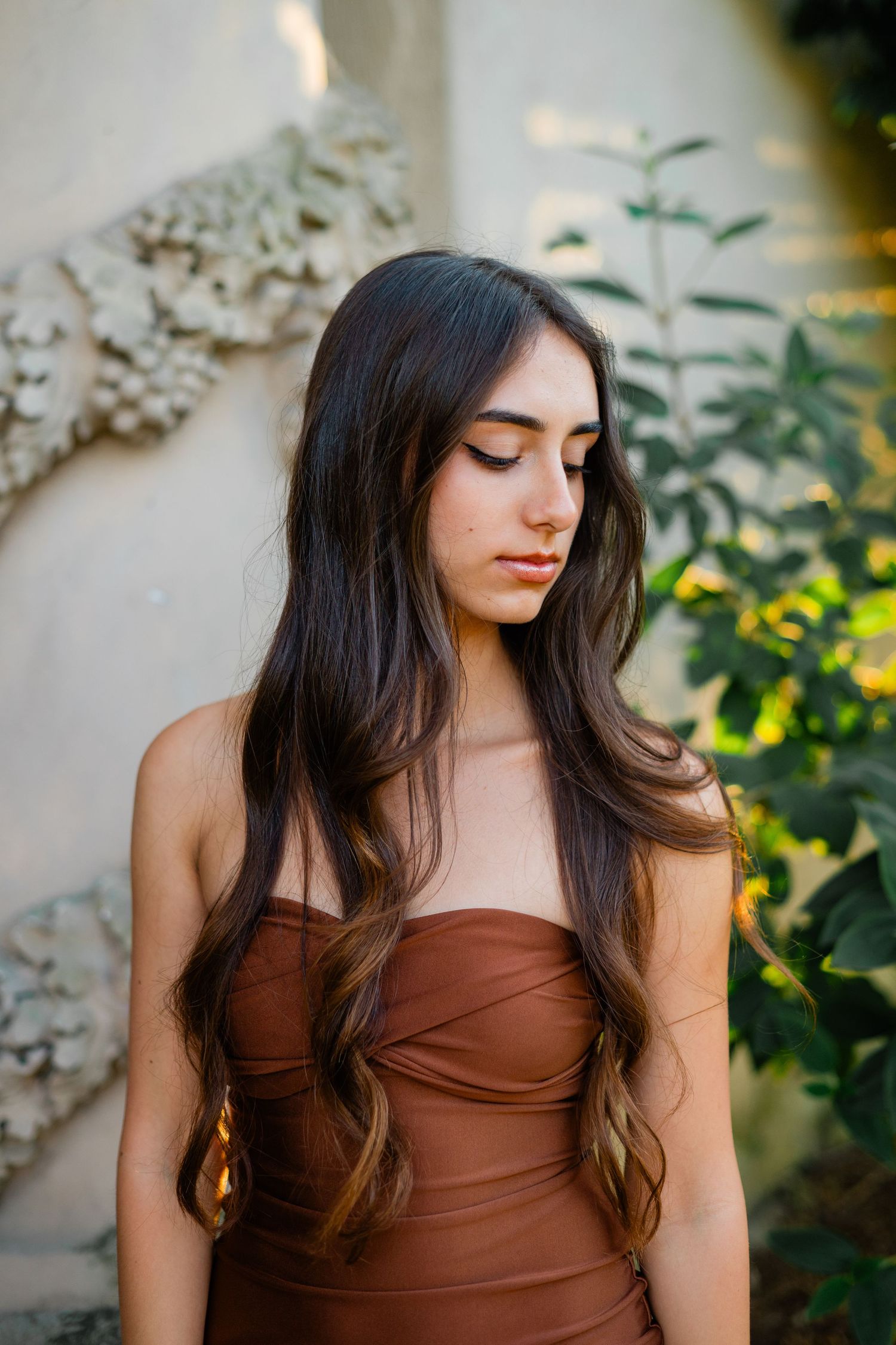 Young woman with long dark hair in brown strapless dress posing outdoors.