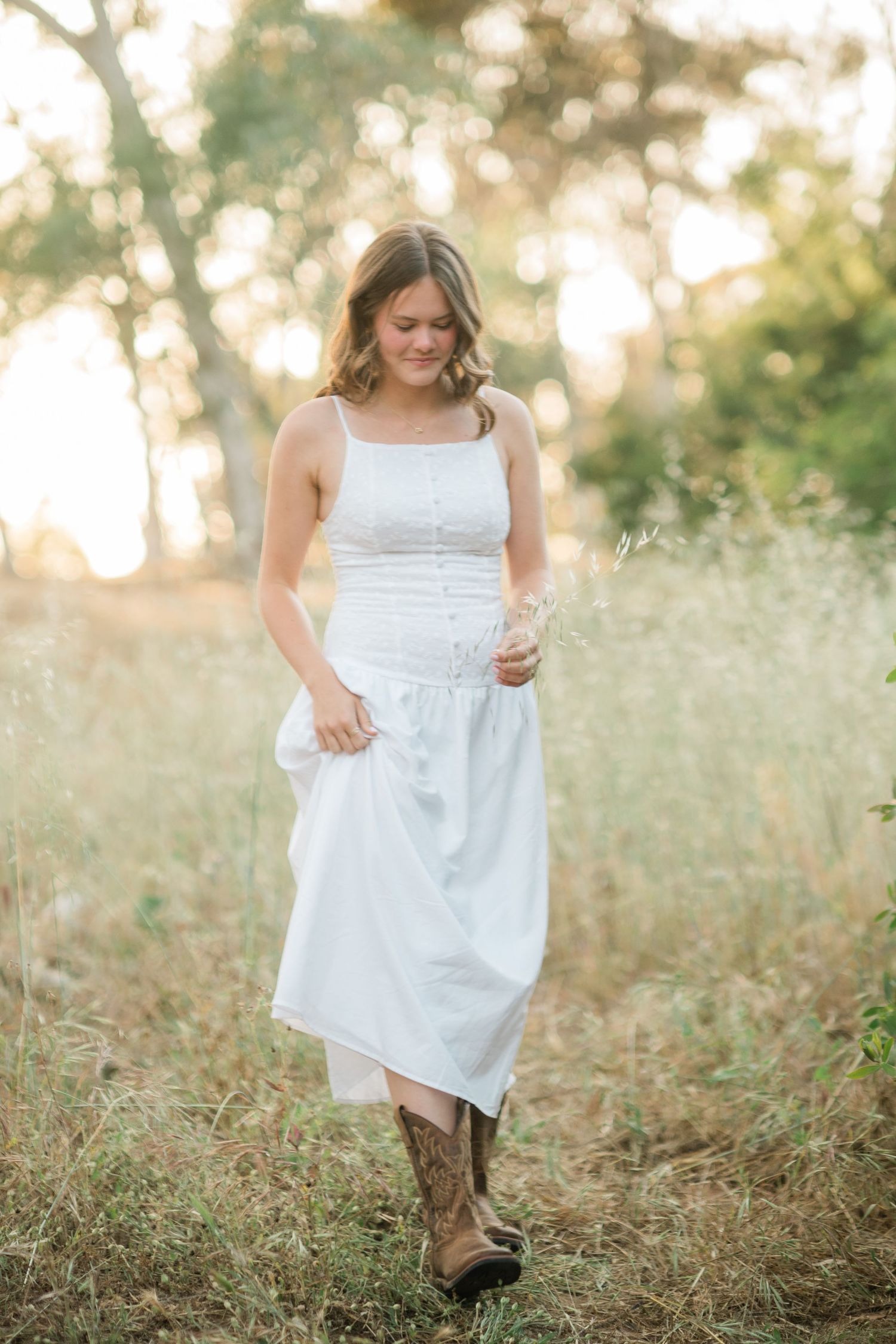 Young woman in white midi dress and cowboy boots walks through golden field holding wildflowers at sunset.