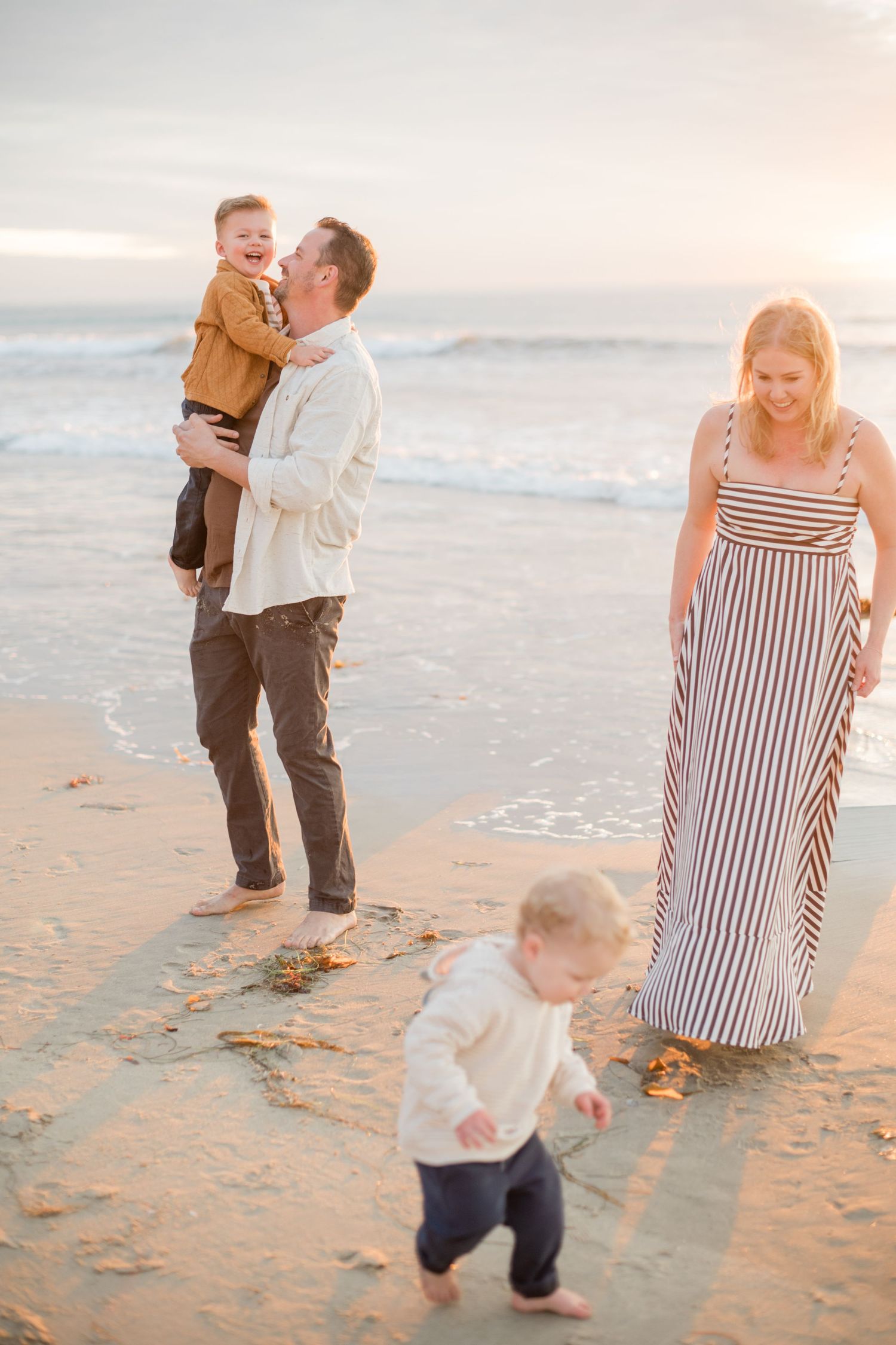 Happy family of four playing on sandy beach at sunset, father holding toddler son.