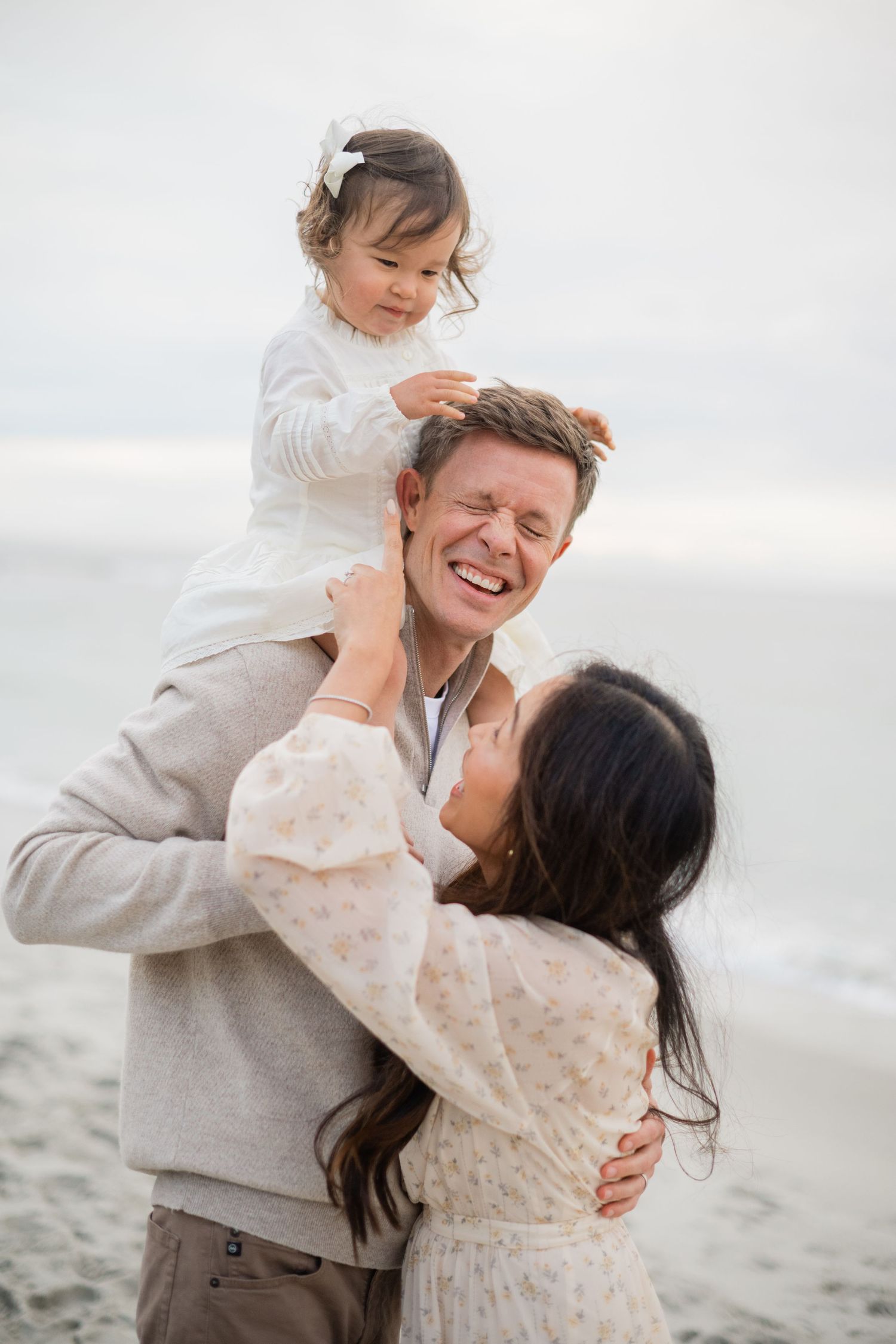 Happy family at the beach, dad holds toddler girl on shoulders while mom embraces them both.