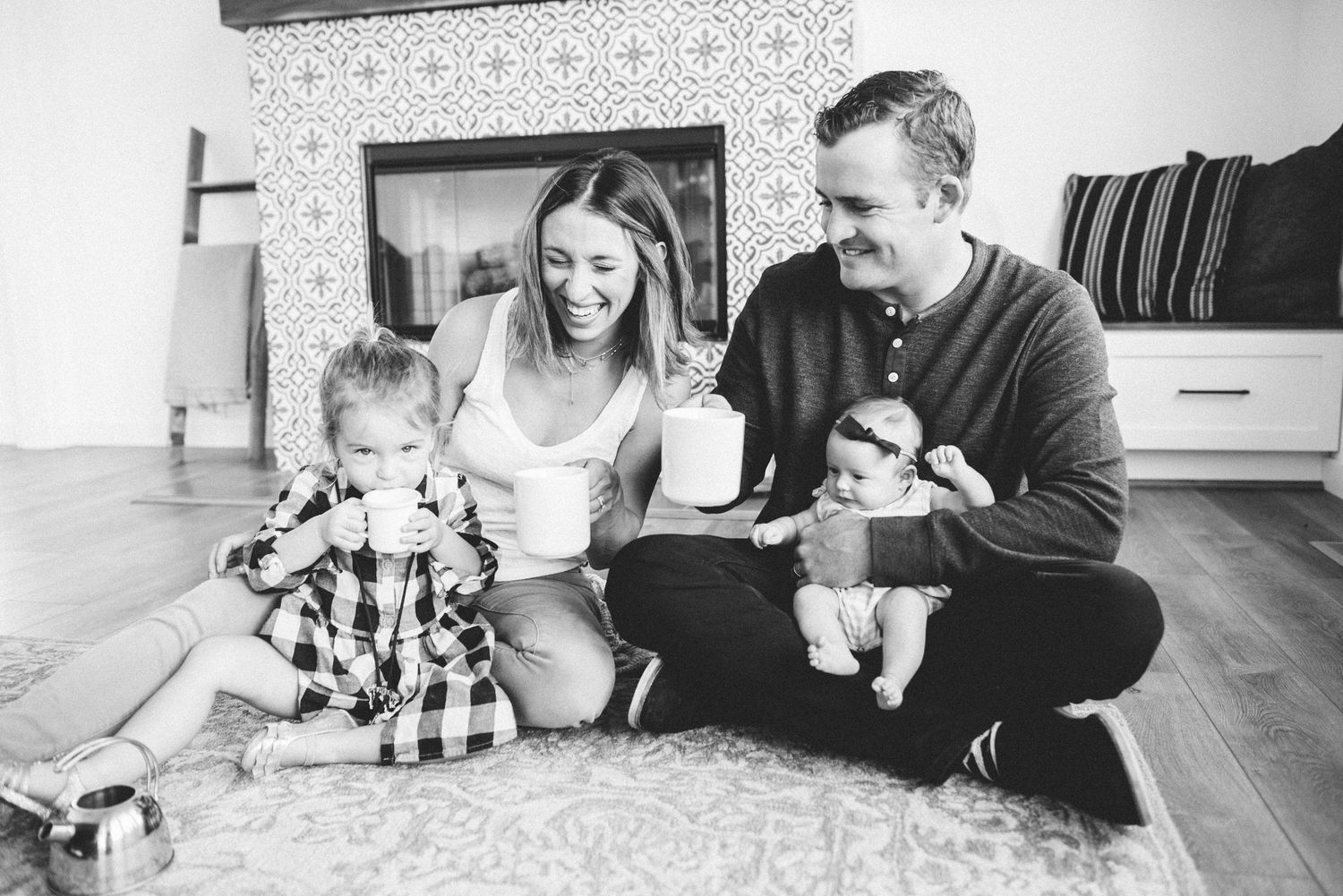 Black and white family photo of parents and two young children sitting on a rug by a fireplace at home.