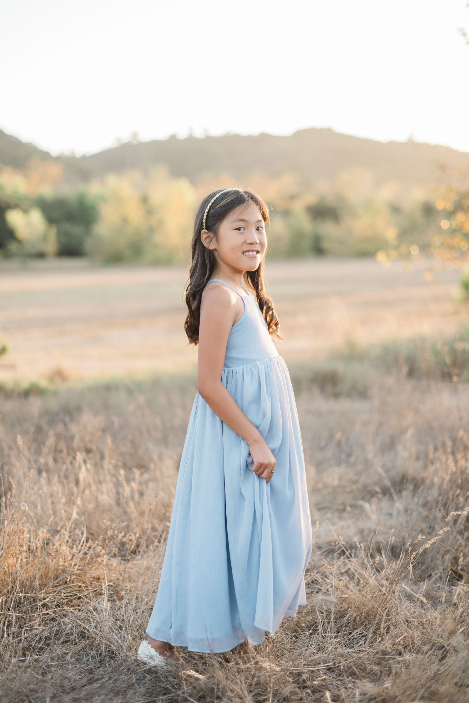 A young girl in a light blue maxi dress smiles in a golden field at sunset.