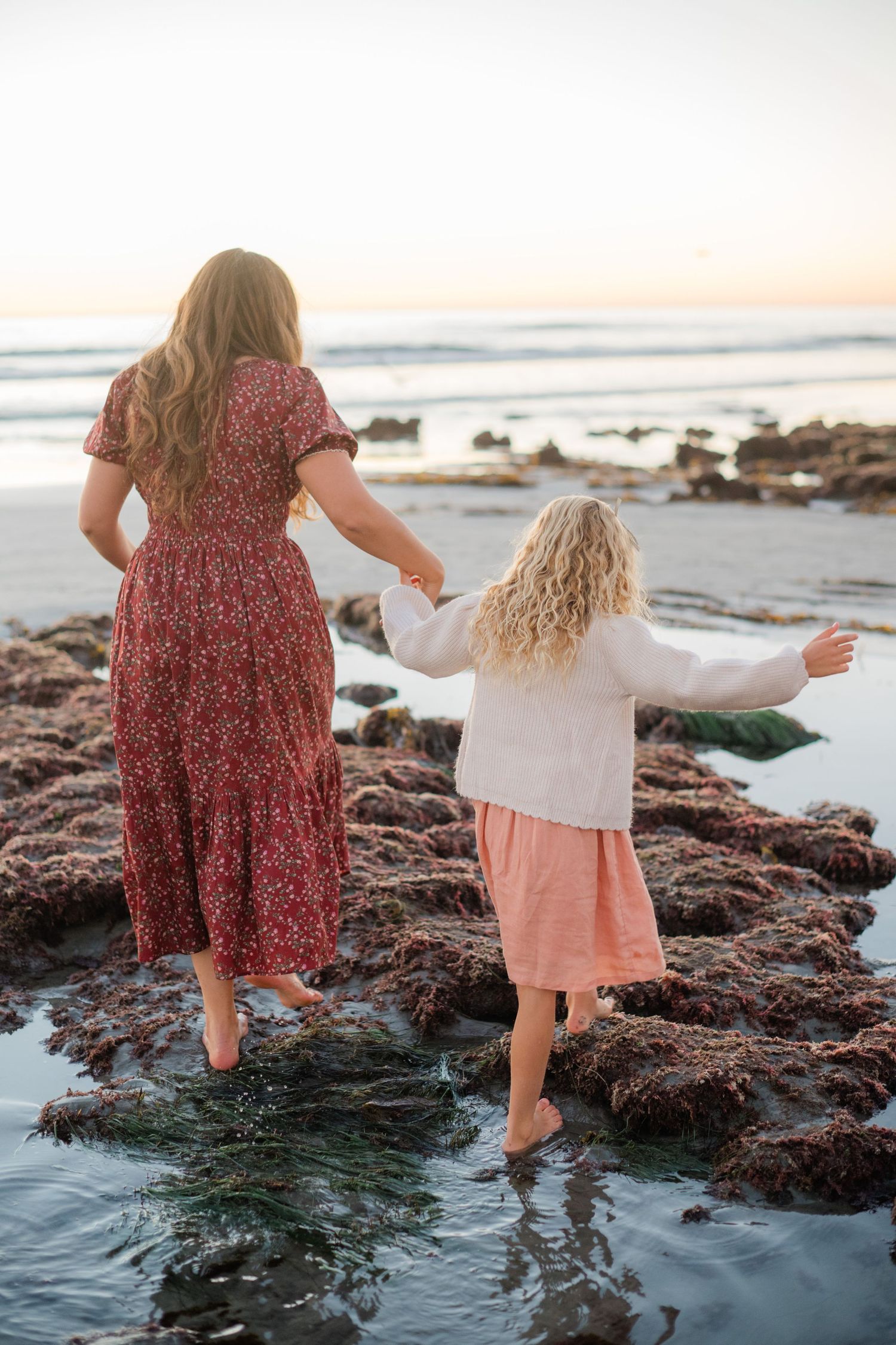 Mother and daughter holding hands while exploring rocky tide pools at the beach during sunset.