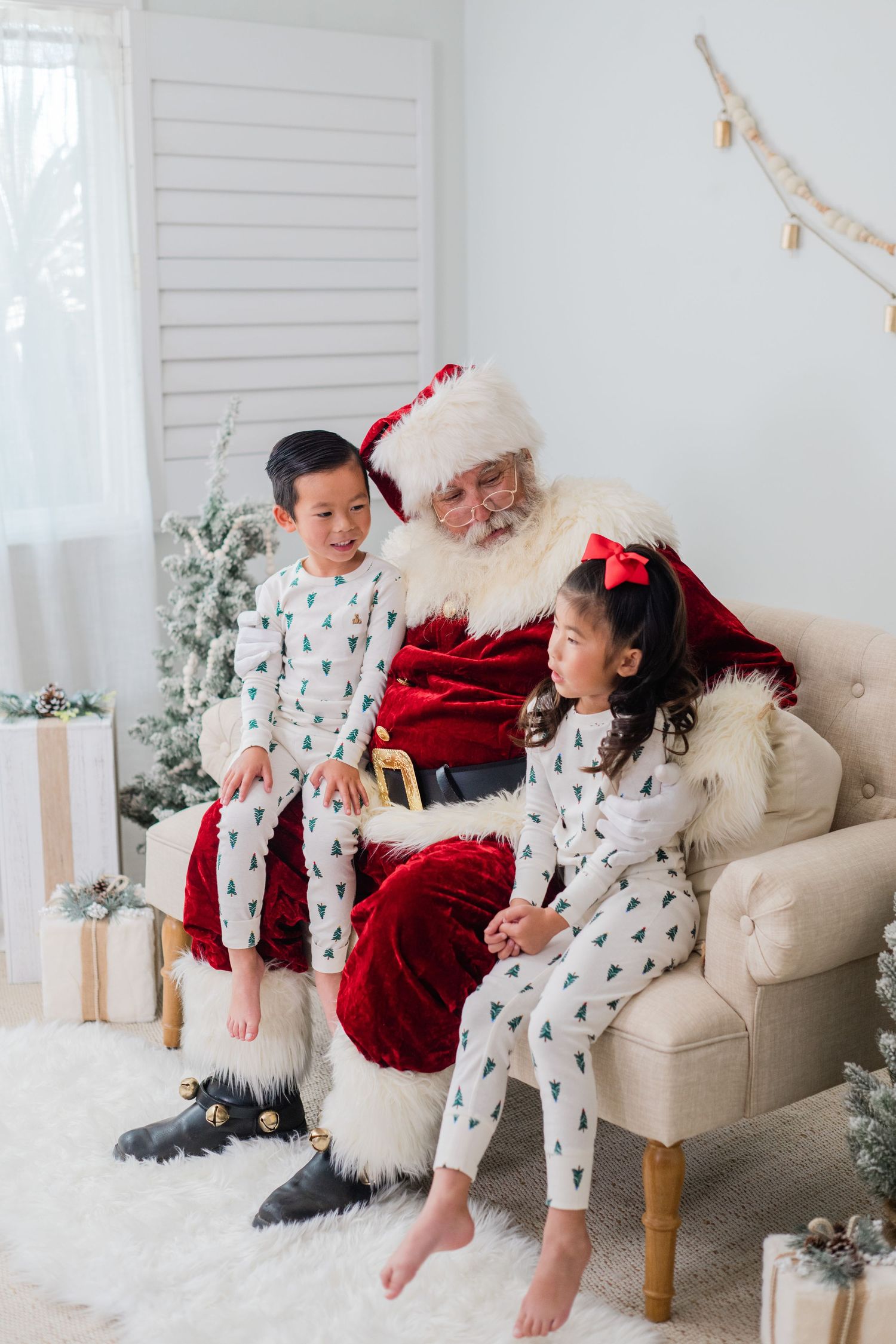 A boy and girl in matching Christmas tree pajamas sit with Santa Claus on a cream armchair in a snowy holiday scene.
