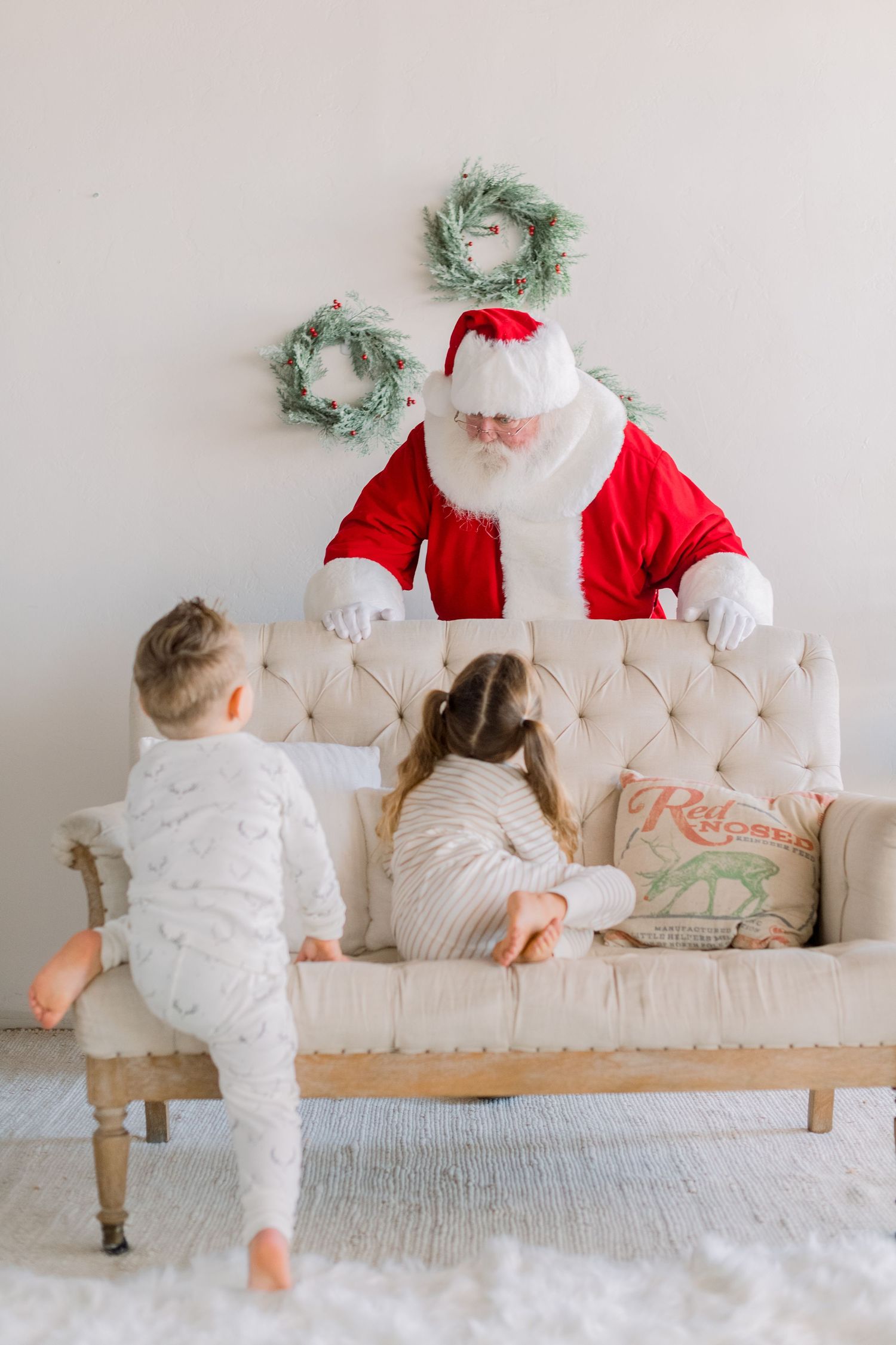 Two young children in pajamas approach Santa Claus sitting on a tufted cream sofa with holiday wreaths on the wall.
