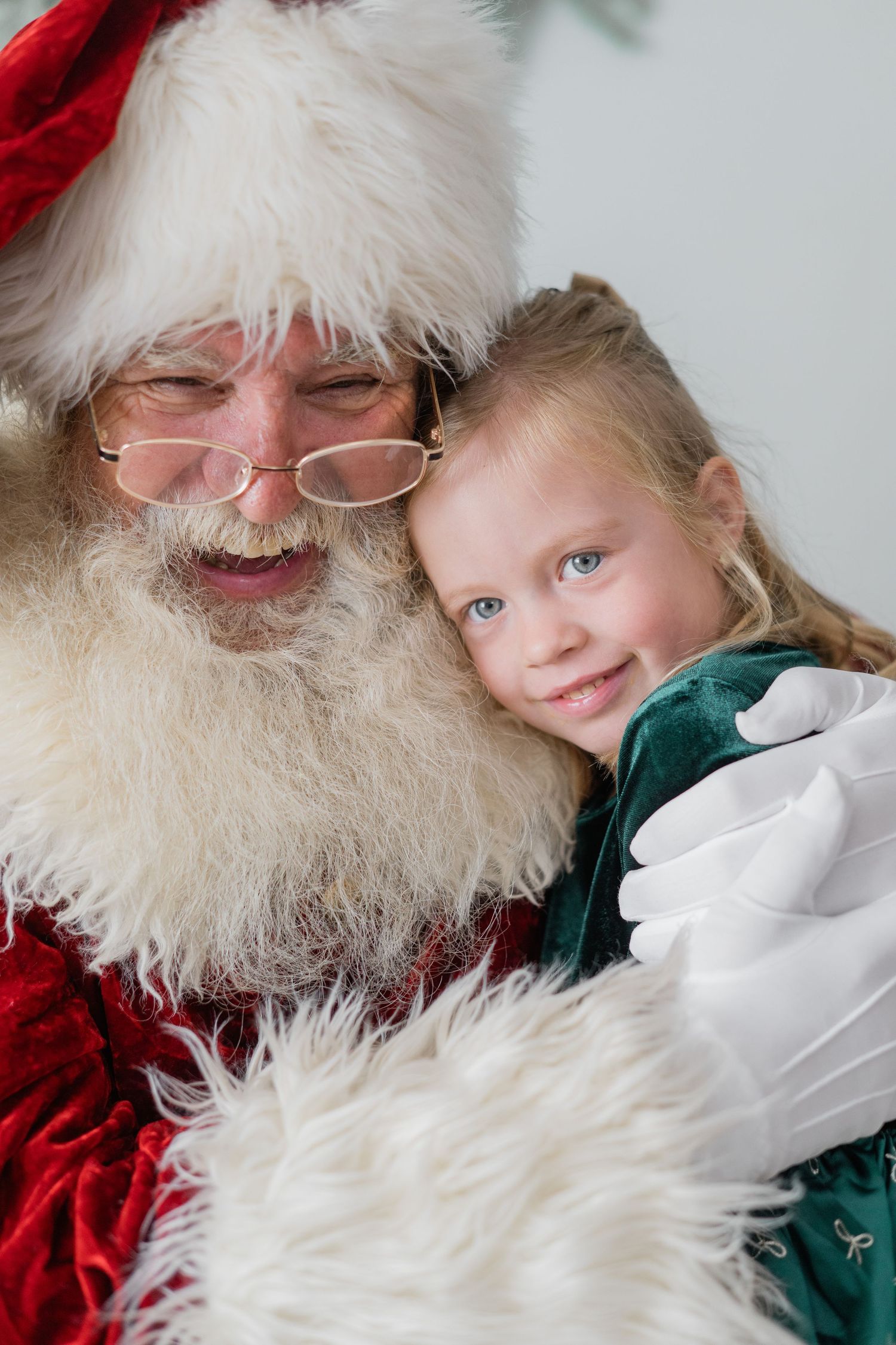 Santa Claus hugging a smiling young blonde girl in a green velvet dress during a Christmas photo.