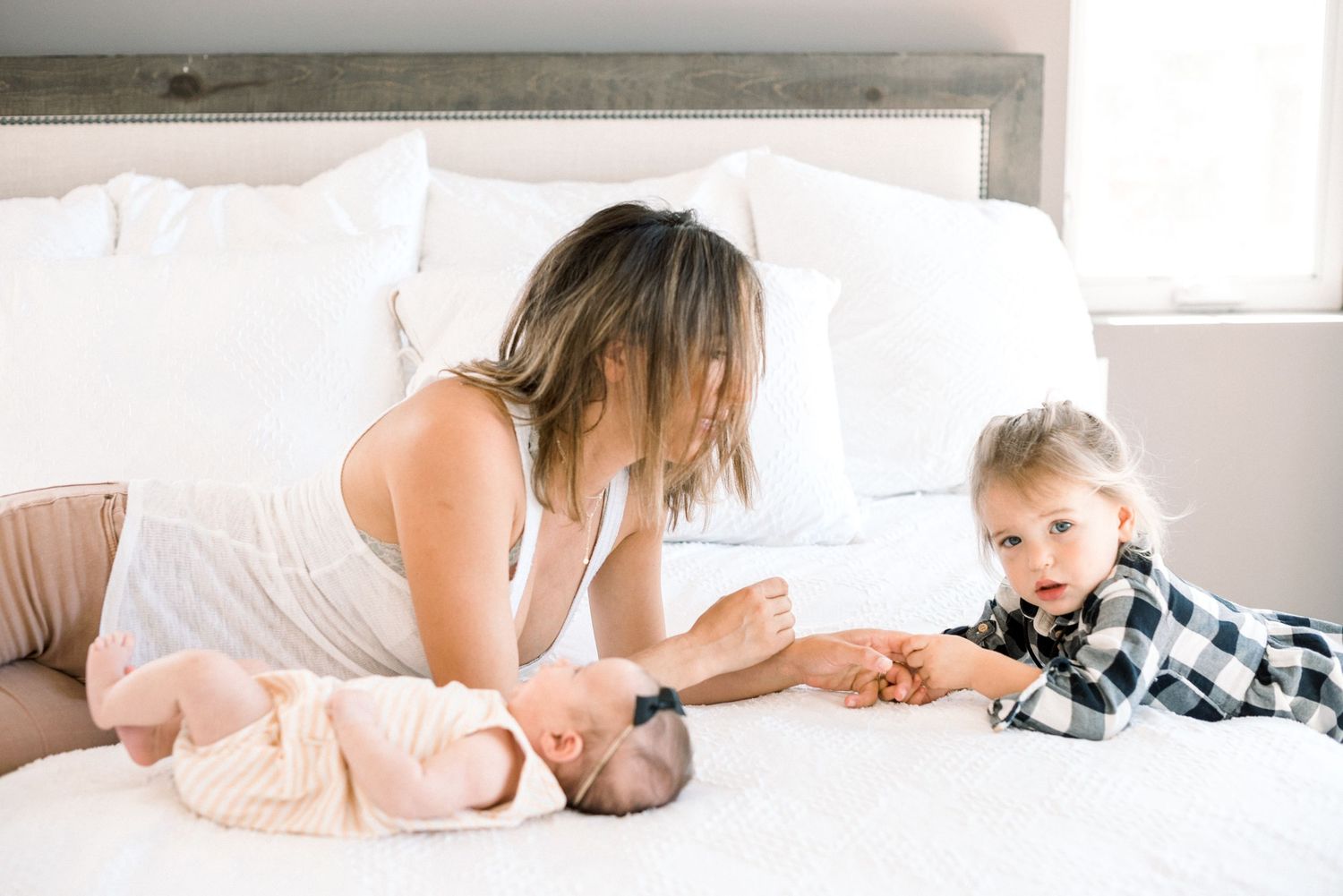 Mother and toddler on white bed admiring swaddled newborn, sibling reaching out to touch baby.