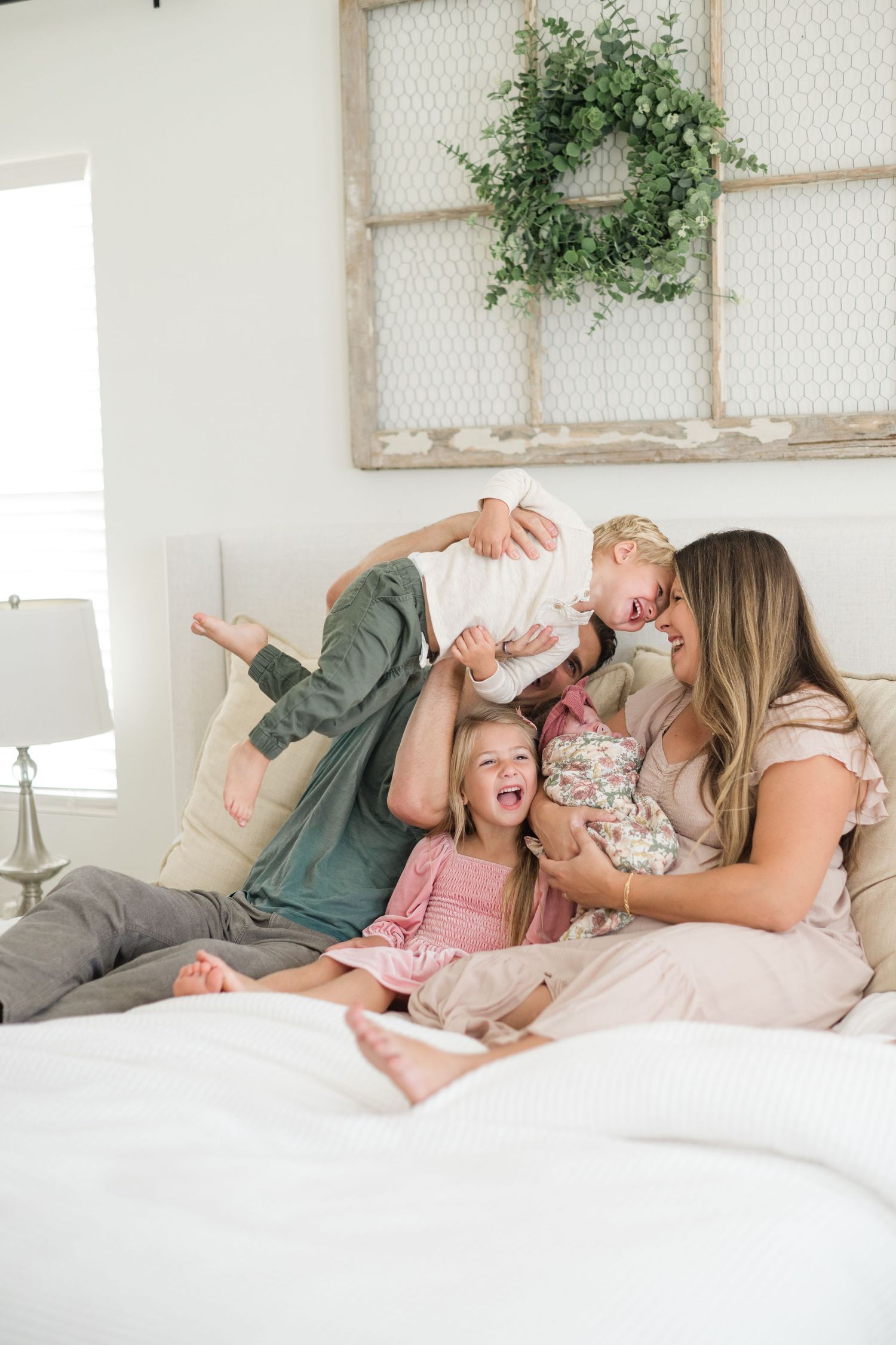 Family laughing and playing together on a white bed in a bright farmhouse-style bedroom with greenery decor.