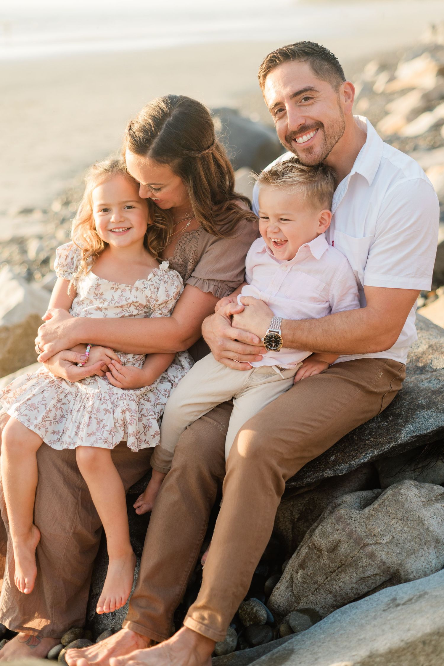 A happy family of four sits on beach rocks, parents embracing their young daughter and son in warm golden light.