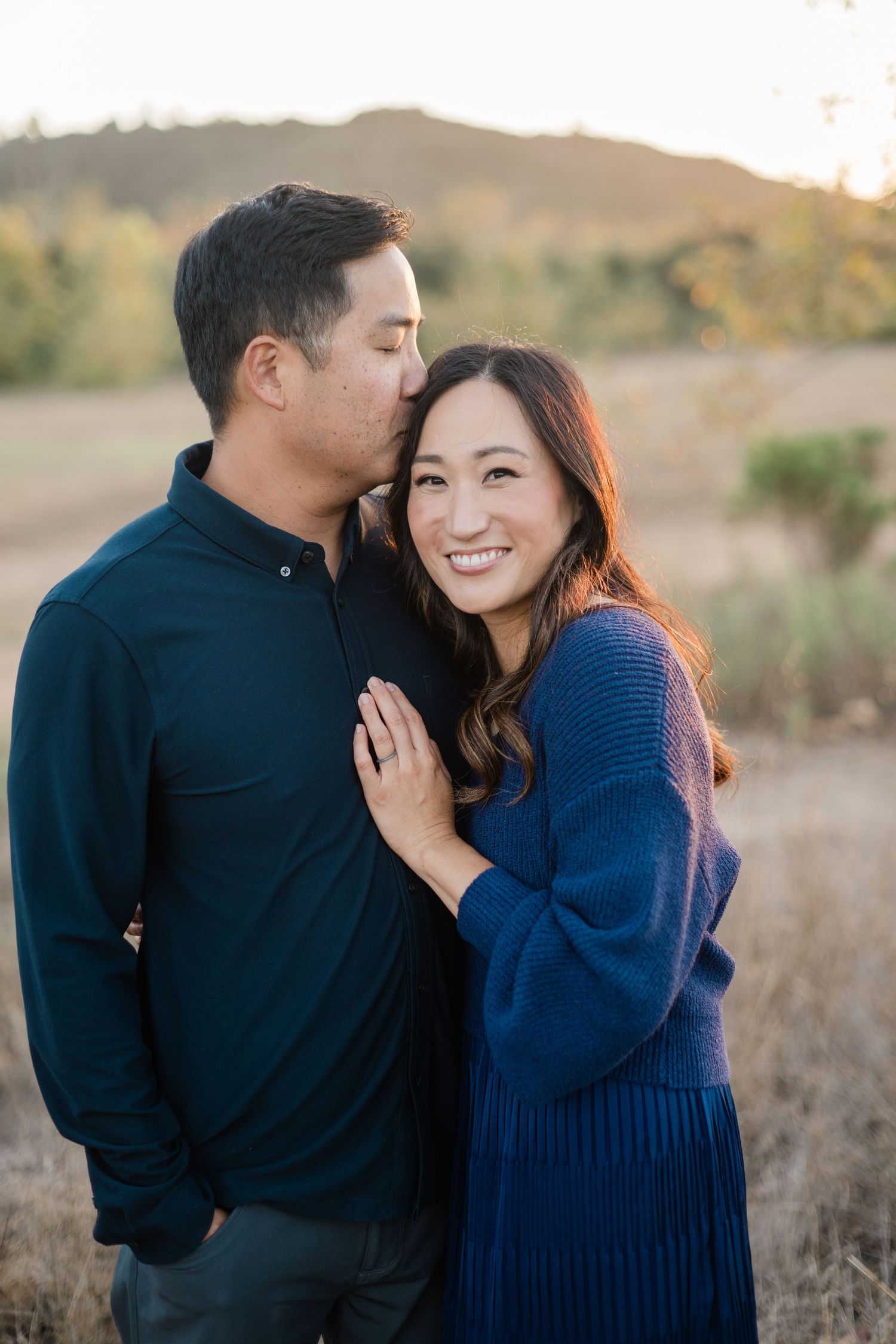 Couple embracing outdoors in golden field, woman in blue sweater smiling, man kissing her cheek, hills in background.