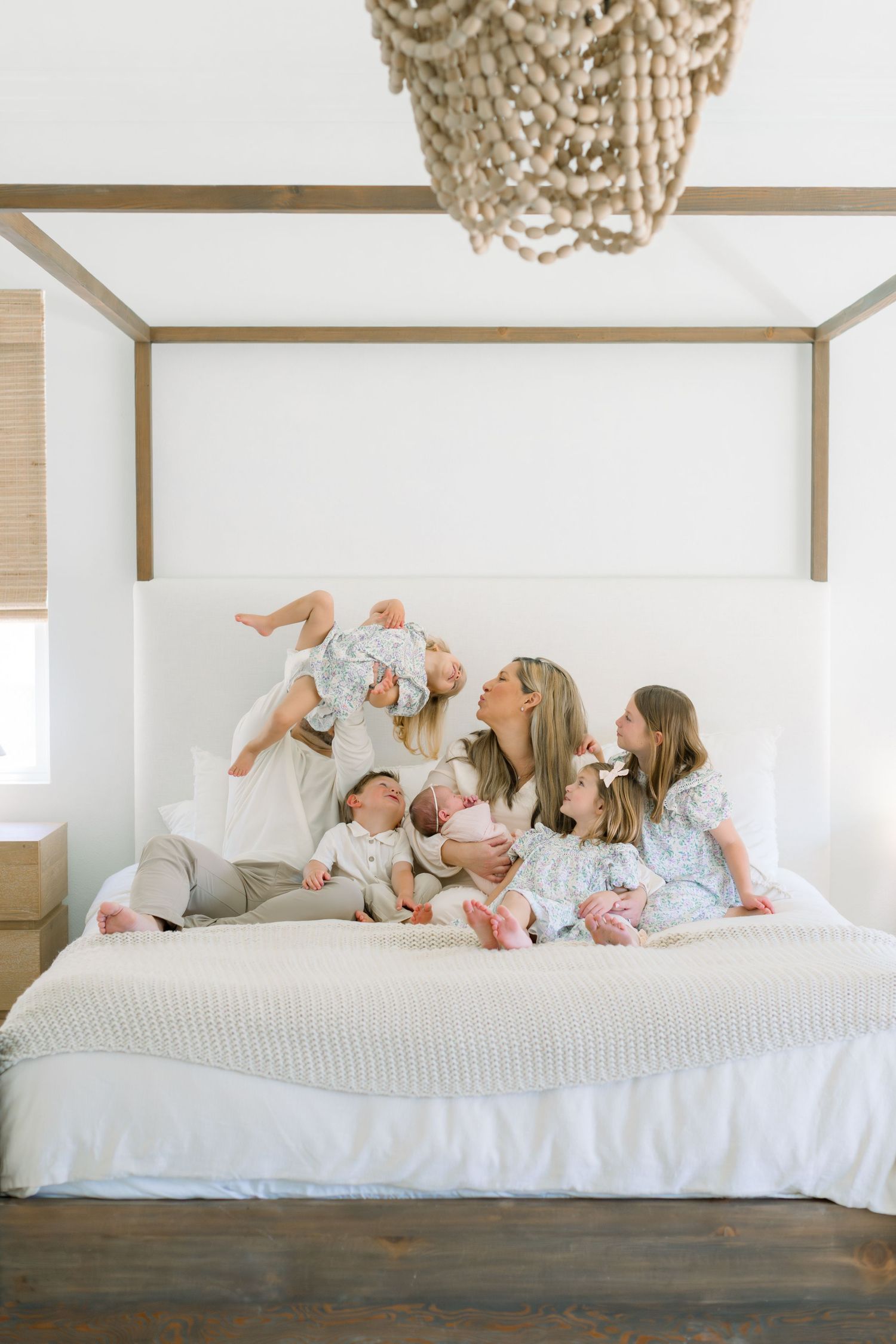 A mother sits on a white canopy bed with four young children in a bright, airy bedroom with a beaded chandelier.