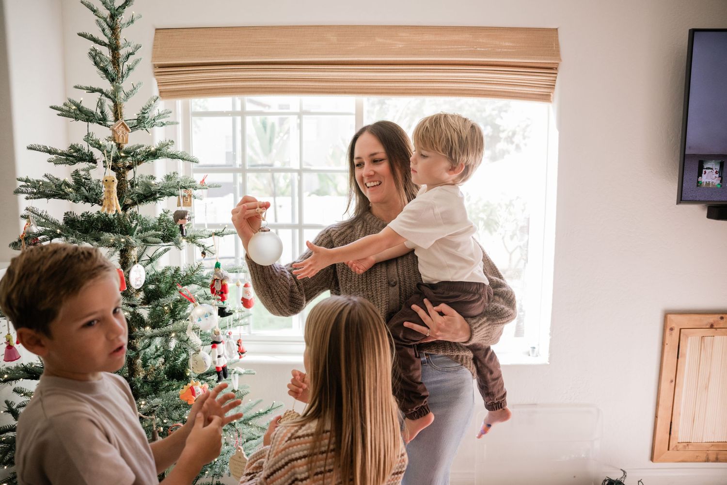 Family decorating a Christmas tree together, mom holding toddler while children hang ornaments in bright living room.