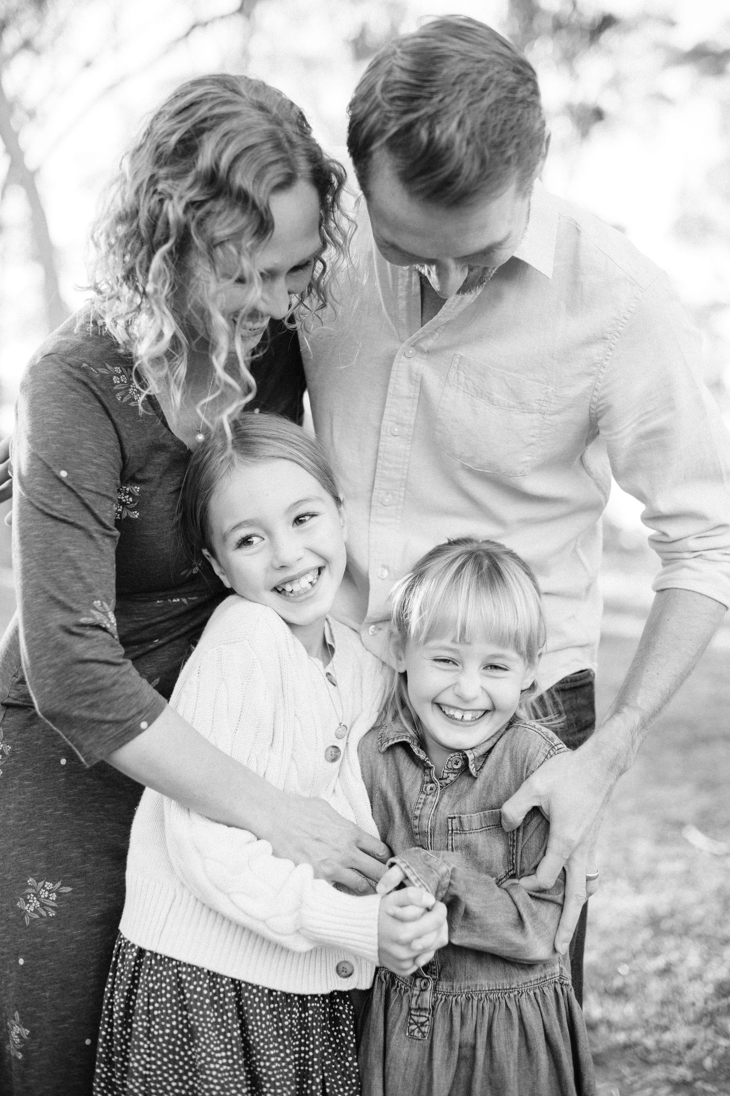 Black and white family portrait of parents with two smiling young girls outdoors among trees.