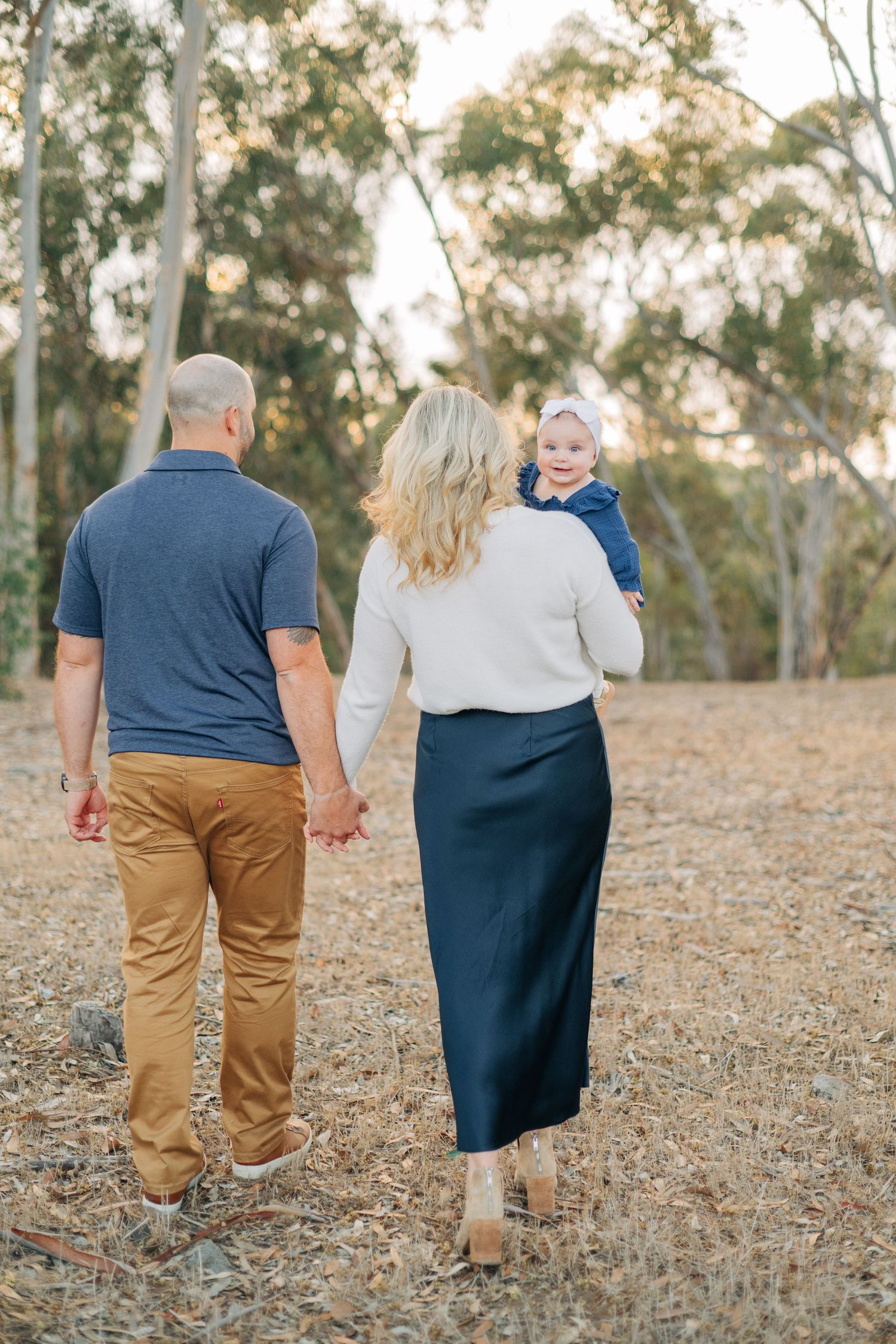 Family of three walking through a sunlit forest, parents holding hands as mother carries baby girl.