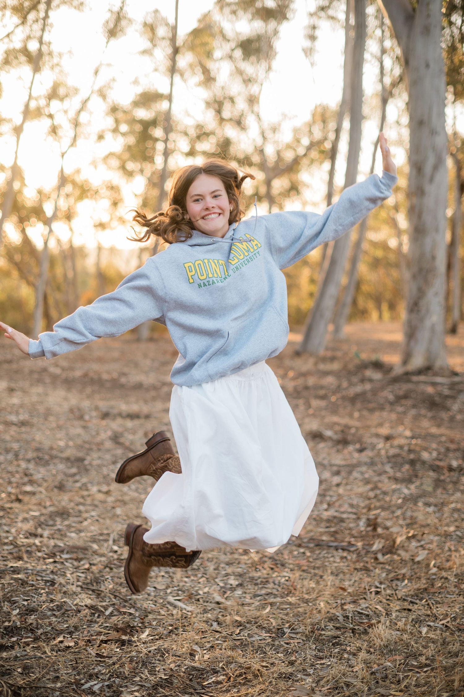 A joyful girl jumps with arms outstretched in a forest, wearing a blue hoodie, white skirt, and brown boots.
