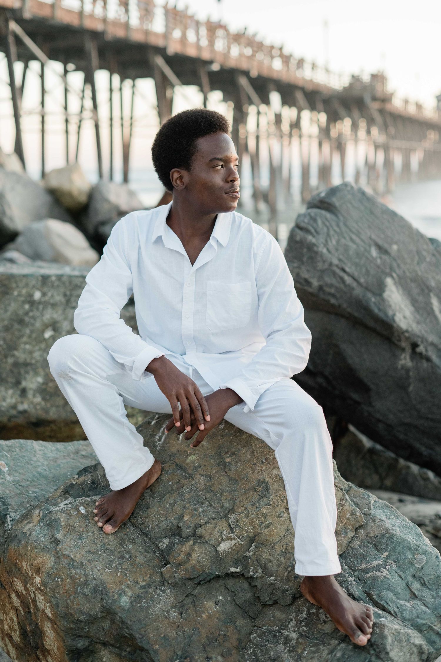 Young Black man in white linen outfit sitting barefoot on rocks near ocean pier at dusk.