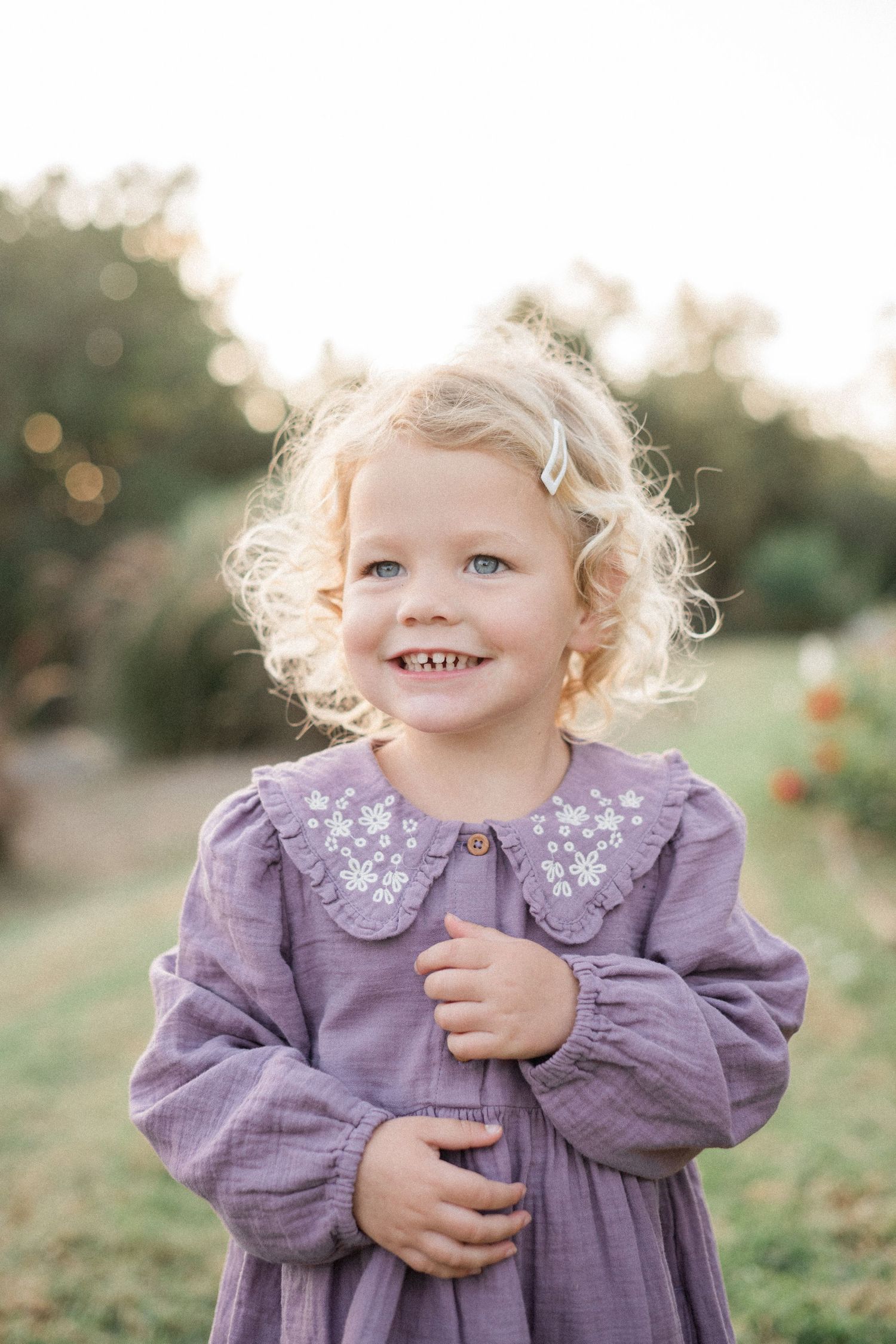 Smiling toddler girl with curly blonde hair wearing a purple floral dress outdoors in soft natural light.