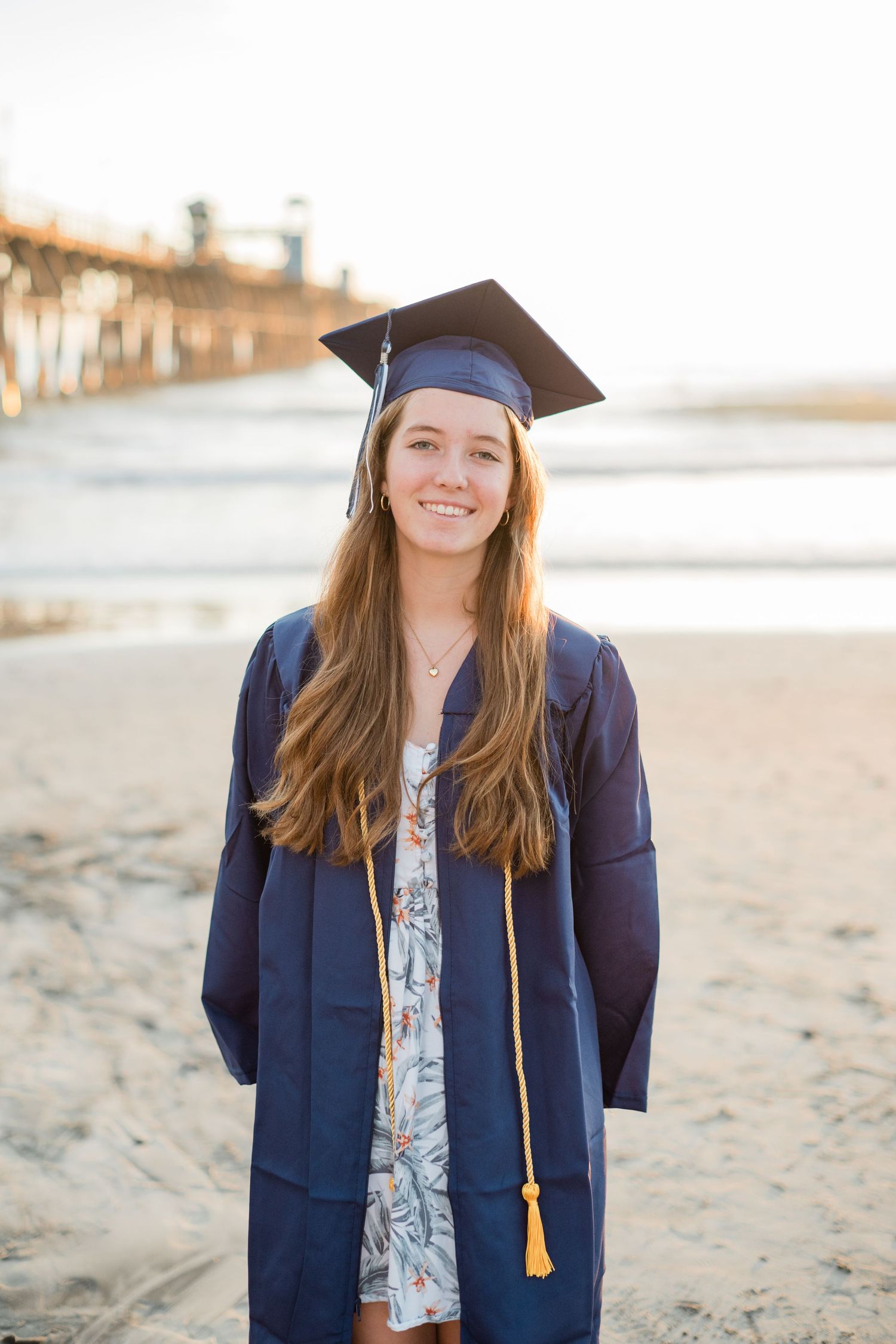 Young woman in navy graduation cap and gown smiling on sandy beach with pier in background at sunset.