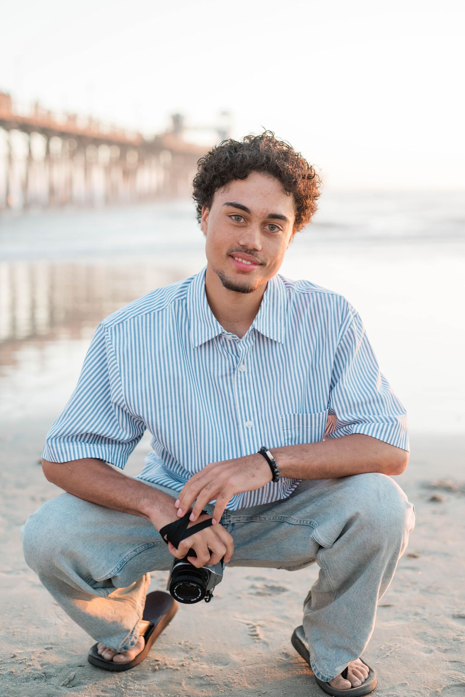 Young man with curly hair crouching on a sandy beach holding a camera, wearing a striped shirt, with a pier in the background.