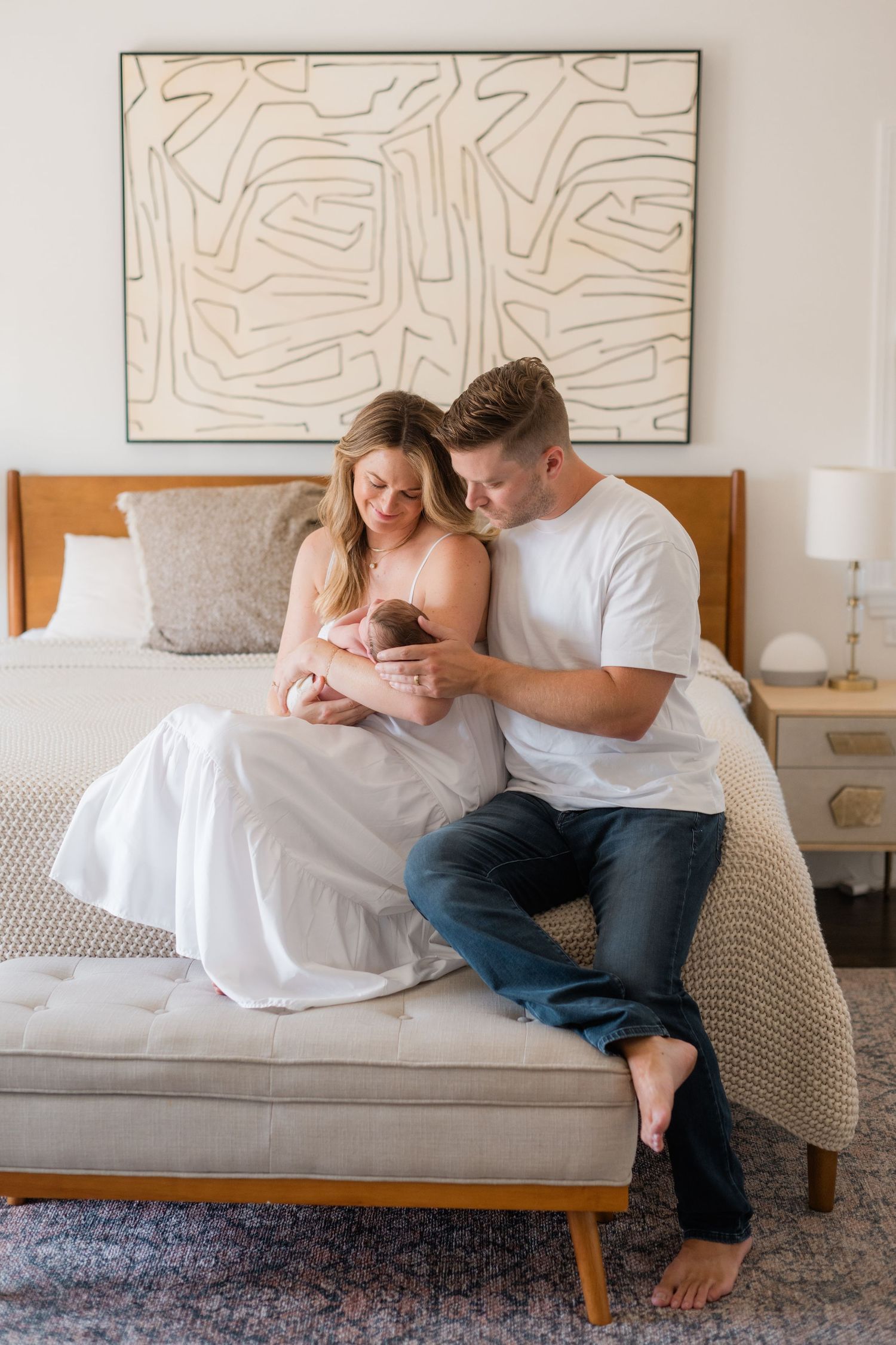 New parents sitting on bed bench lovingly gazing at their swaddled newborn baby.