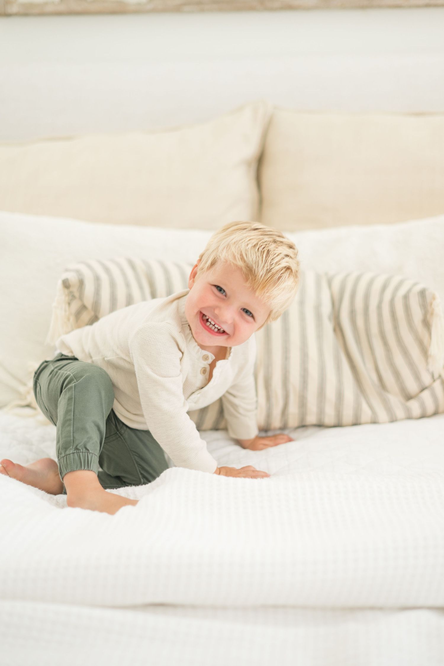 A cheerful blonde toddler boy smiling and playing on a white bed with striped pillows, wearing green pants and cream top.
