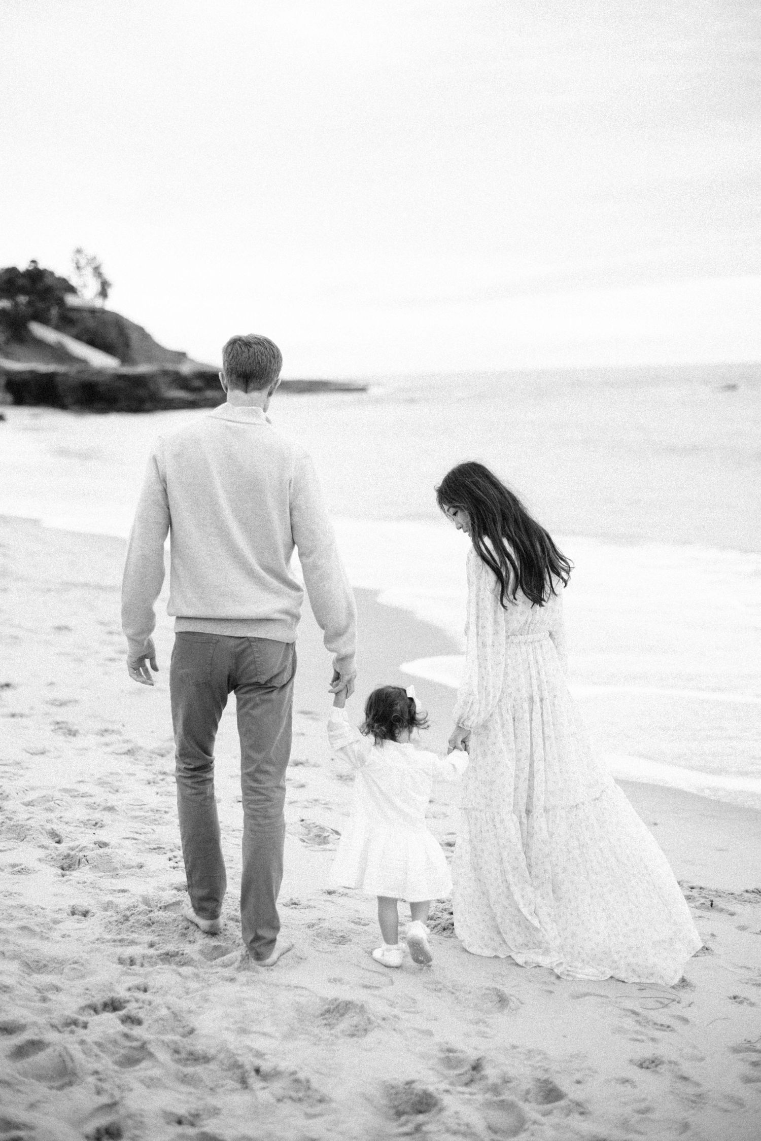 Family of three walks hand-in-hand along sandy beach in black and white photo.