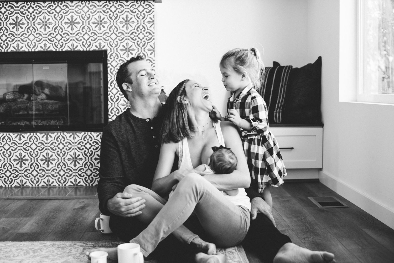 Black and white photo of a joyful family sitting on the floor near a fireplace with a newborn and toddler.