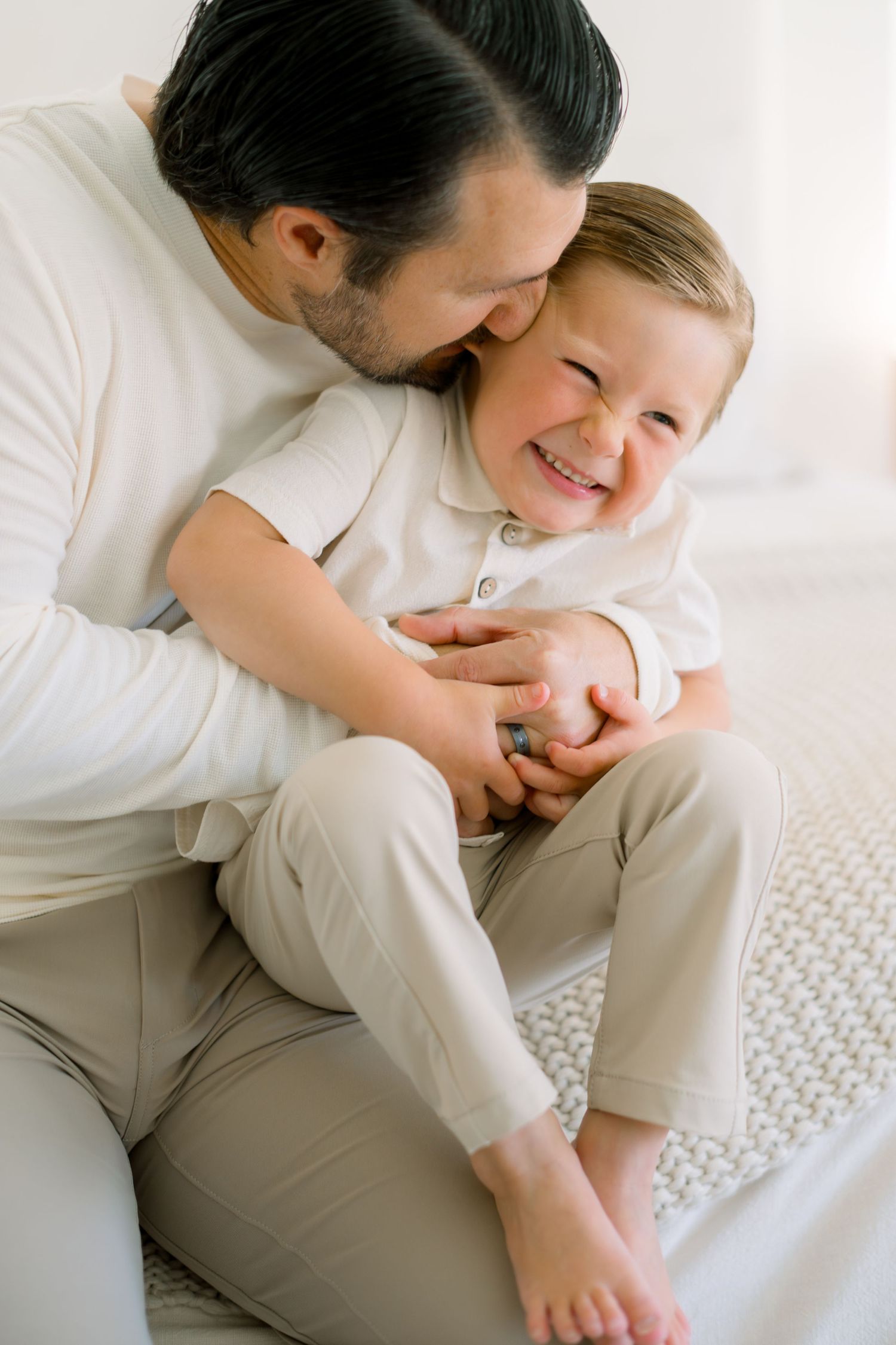 A father kisses his laughing toddler son while sitting together on a bed, both wearing matching cream outfits.