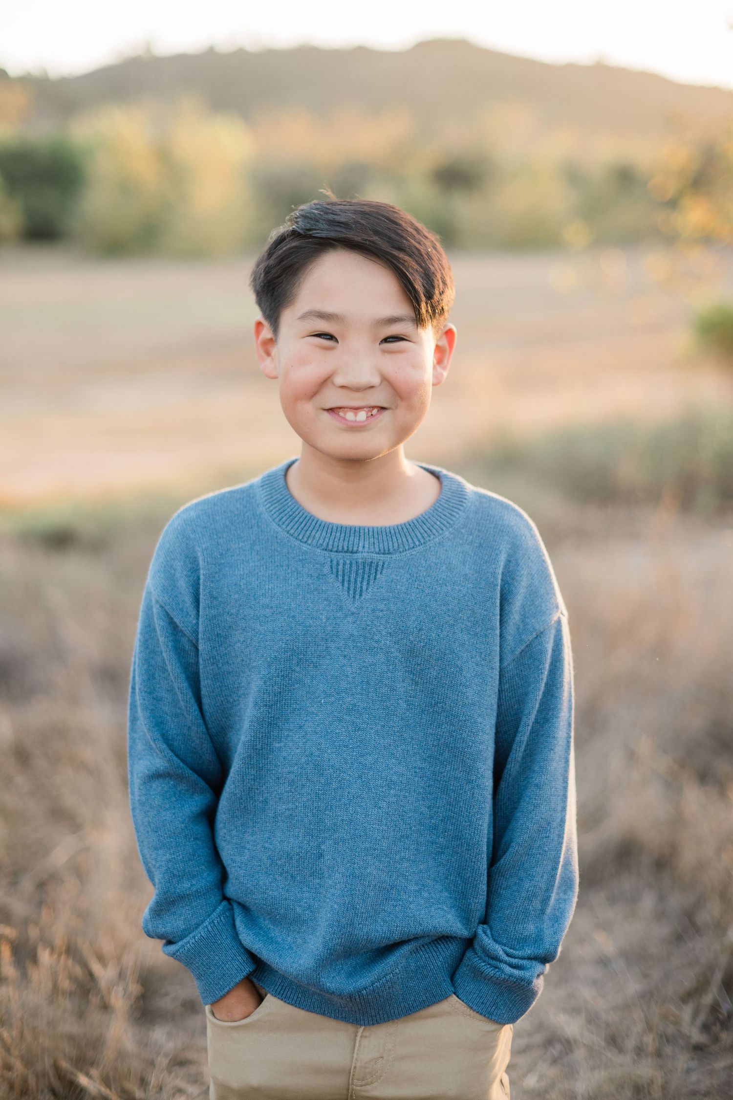 Smiling young boy in a blue sweater standing in a golden field with hills in the background.