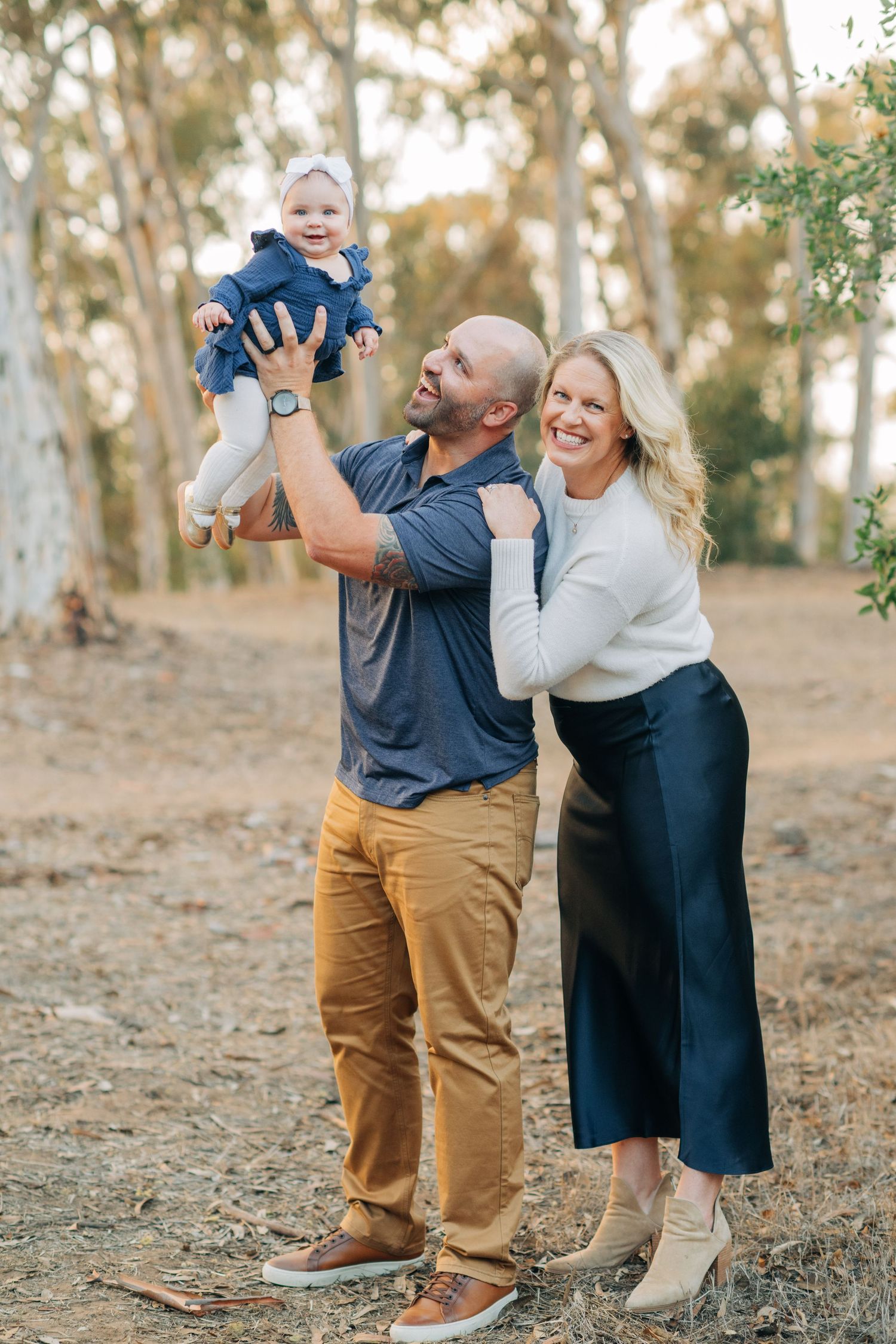 A happy family with a bald dad lifting a smiling baby girl while mom stands beside them outdoors.
