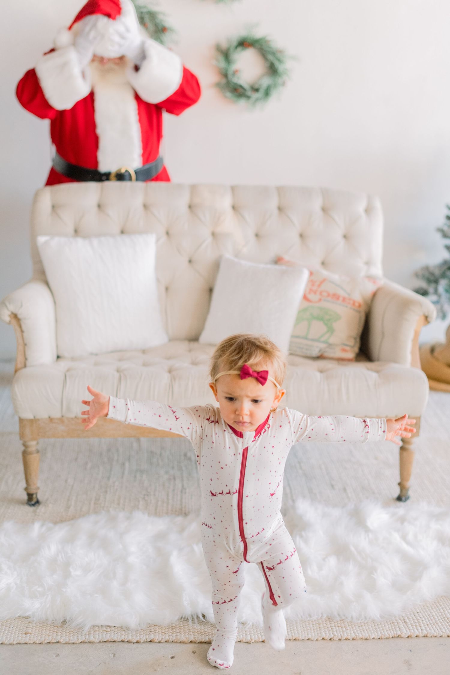 Toddler girl in Christmas pajamas stands with arms outstretched on white rug, Santa Claus visible behind cream tufted sofa.