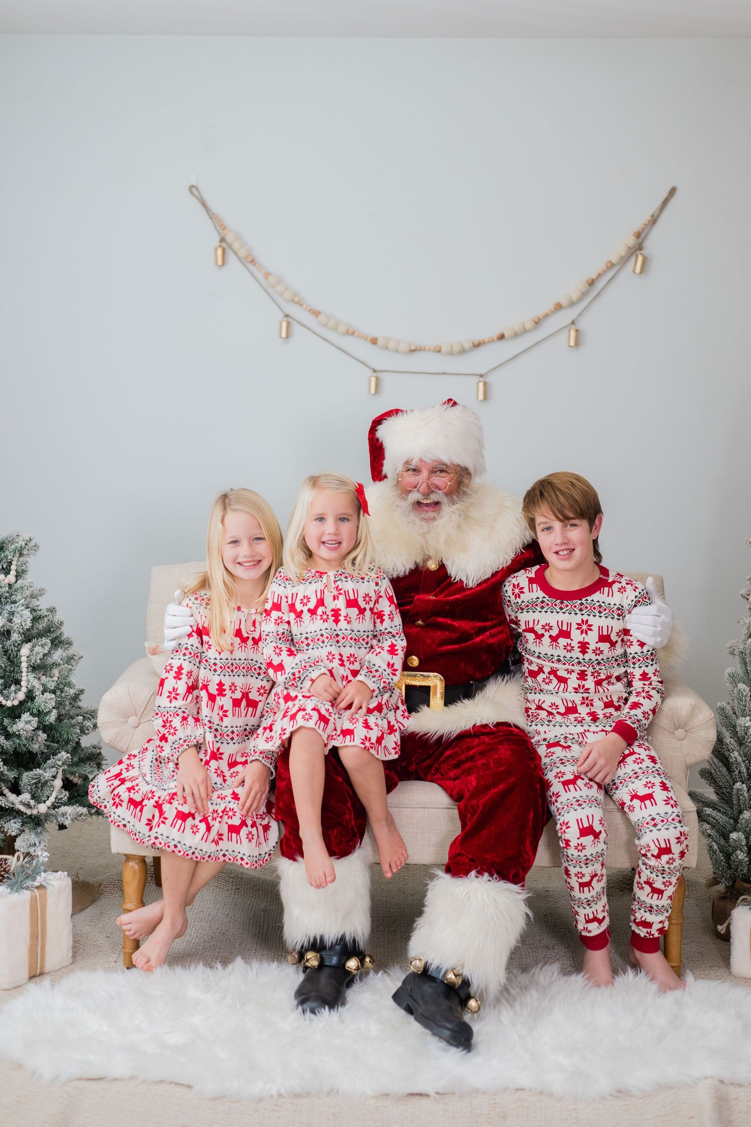 Three children in Christmas pajamas sit smiling with Santa Claus on a couch in a festive holiday photo setting.