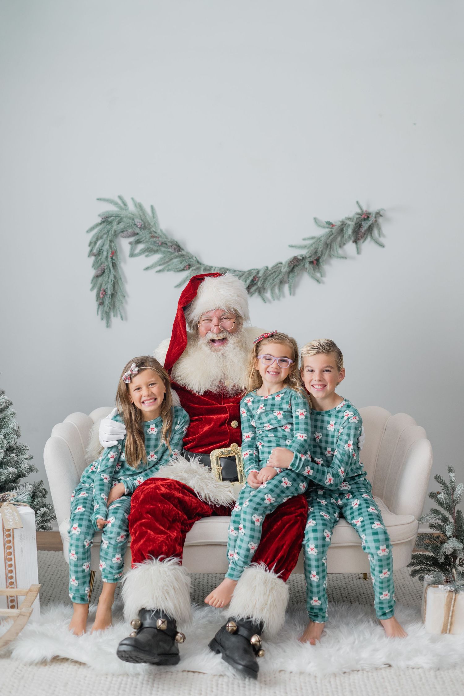 Three children in matching teal pajamas smile with Santa Claus on a white sofa surrounded by festive Christmas decor.