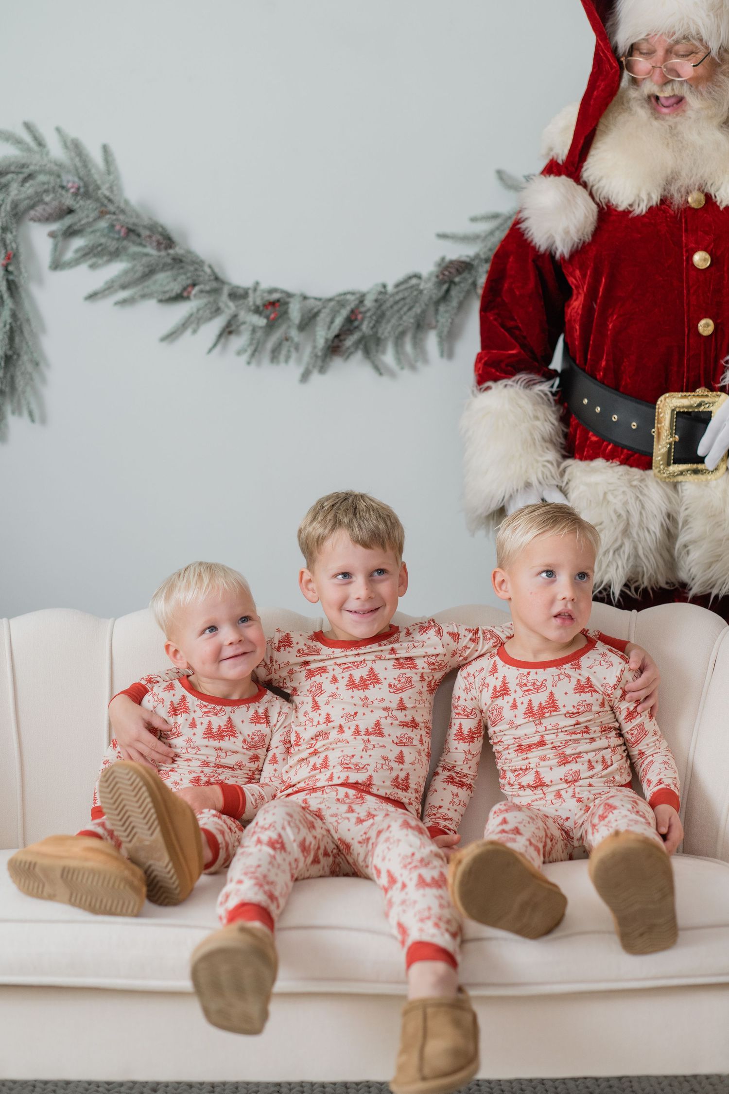Three blonde boys in matching Christmas pajamas sit on a white sofa with Santa Claus behind them.