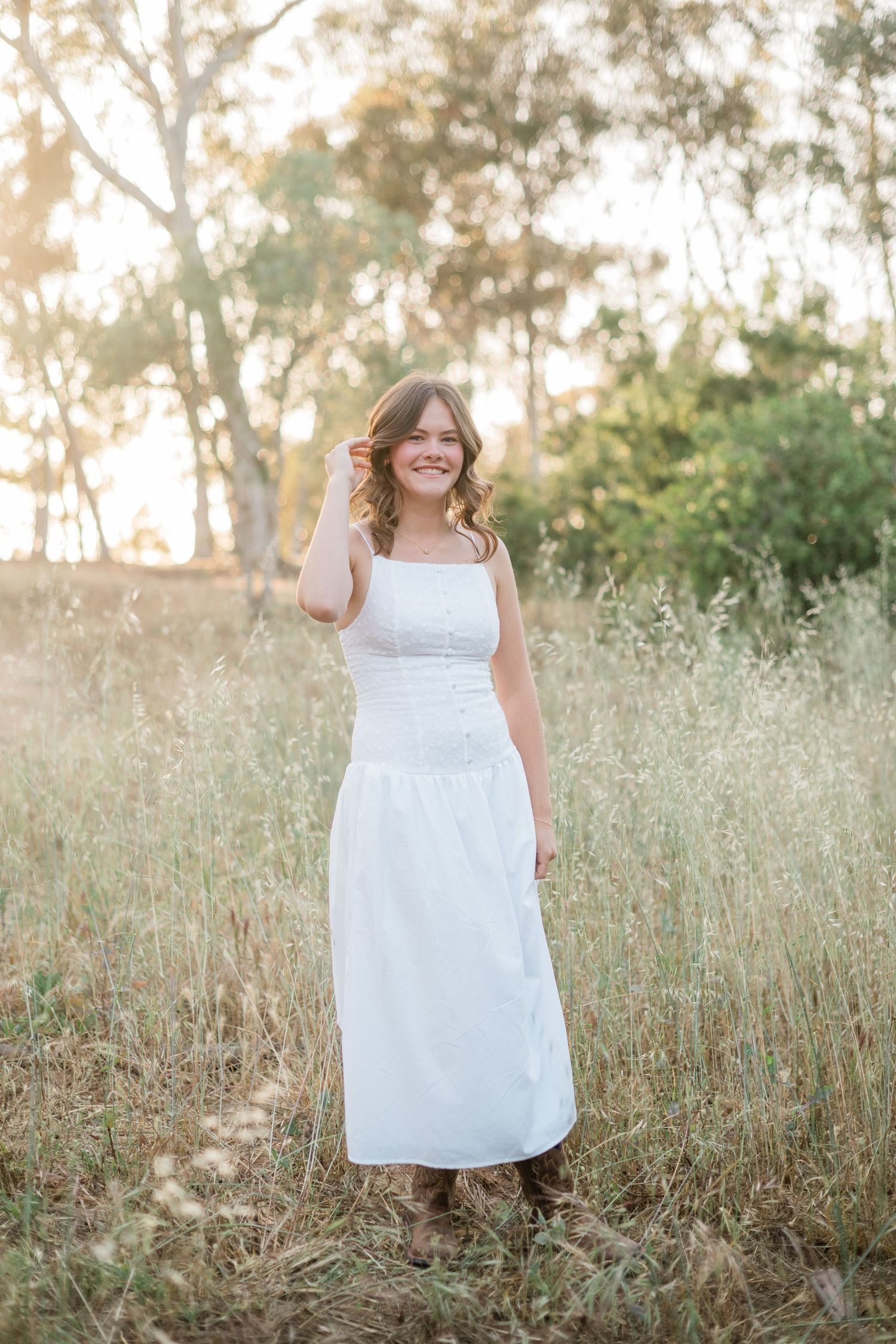 Young woman in white maxi dress and brown boots smiling in golden field with trees in background.