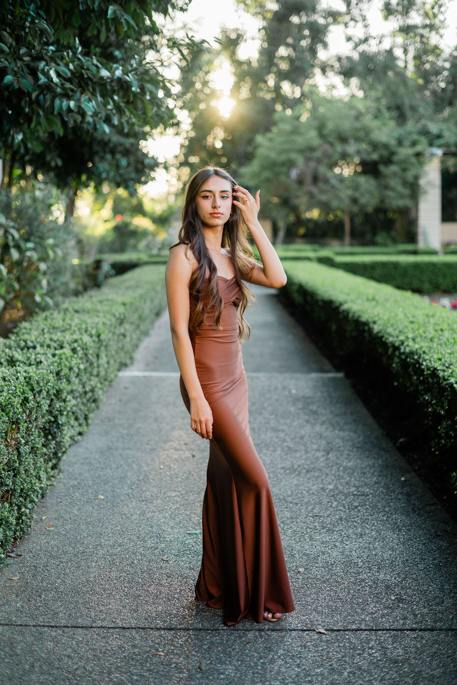 Woman in elegant brown maxi dress posing in a formal garden pathway with lush hedges and golden sunlight.