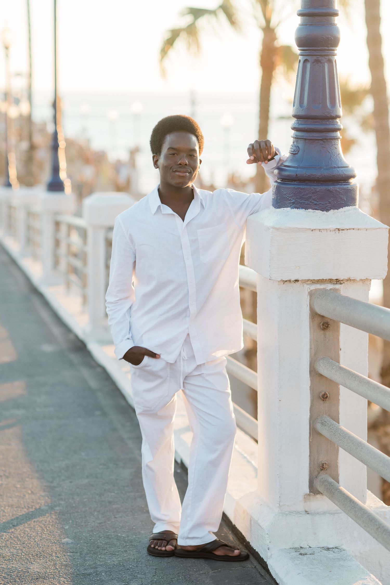 Young Black man in white outfit leaning on railing of seaside promenade with palm trees at sunset.