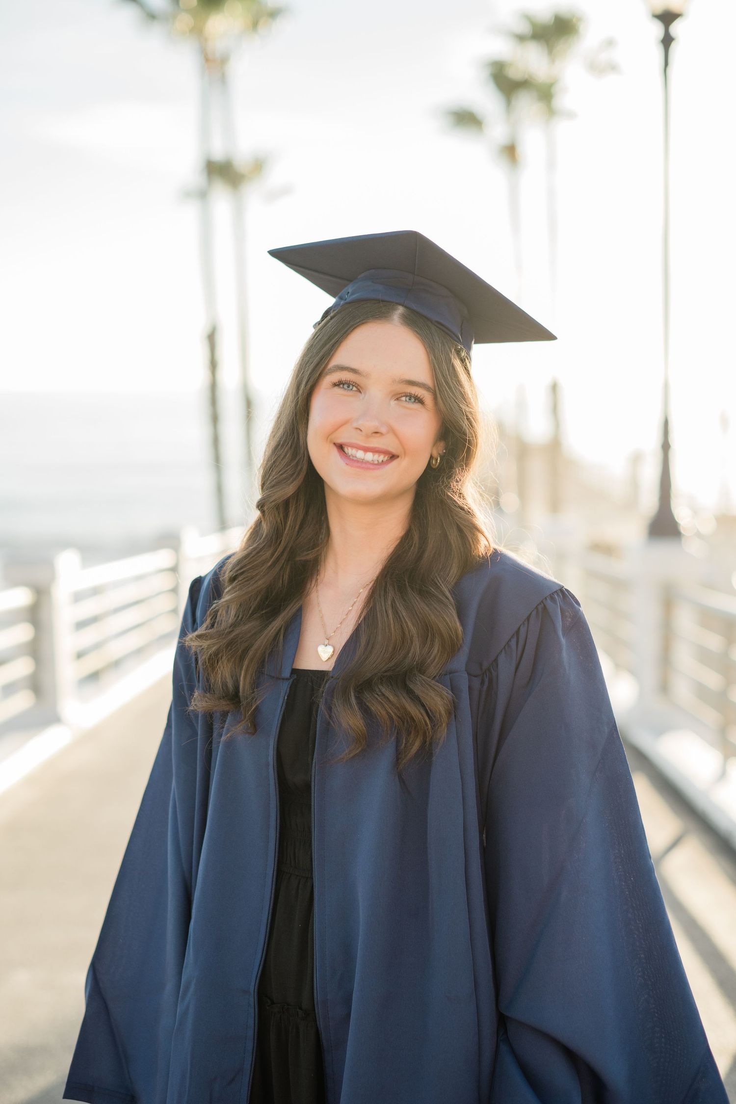 Smiling young woman in navy blue graduation cap and gown posing on a sunny pier with palm trees.