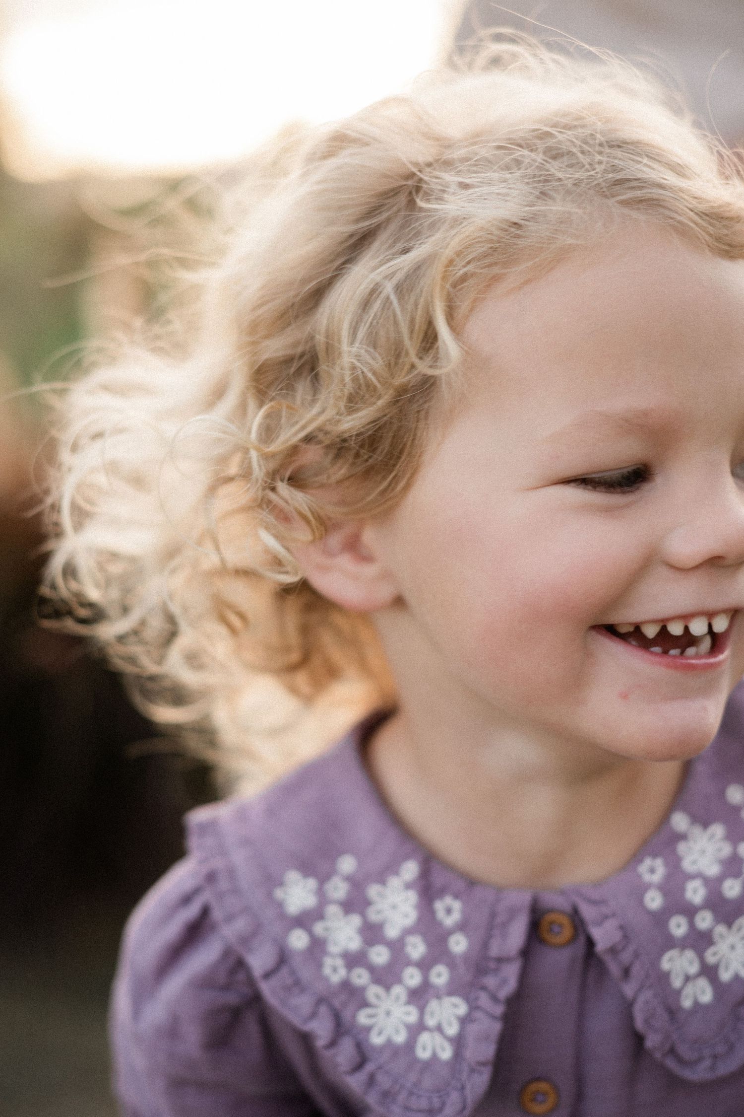 Adorable blonde curly-haired toddler girl laughing in a purple floral dress in golden sunlight.