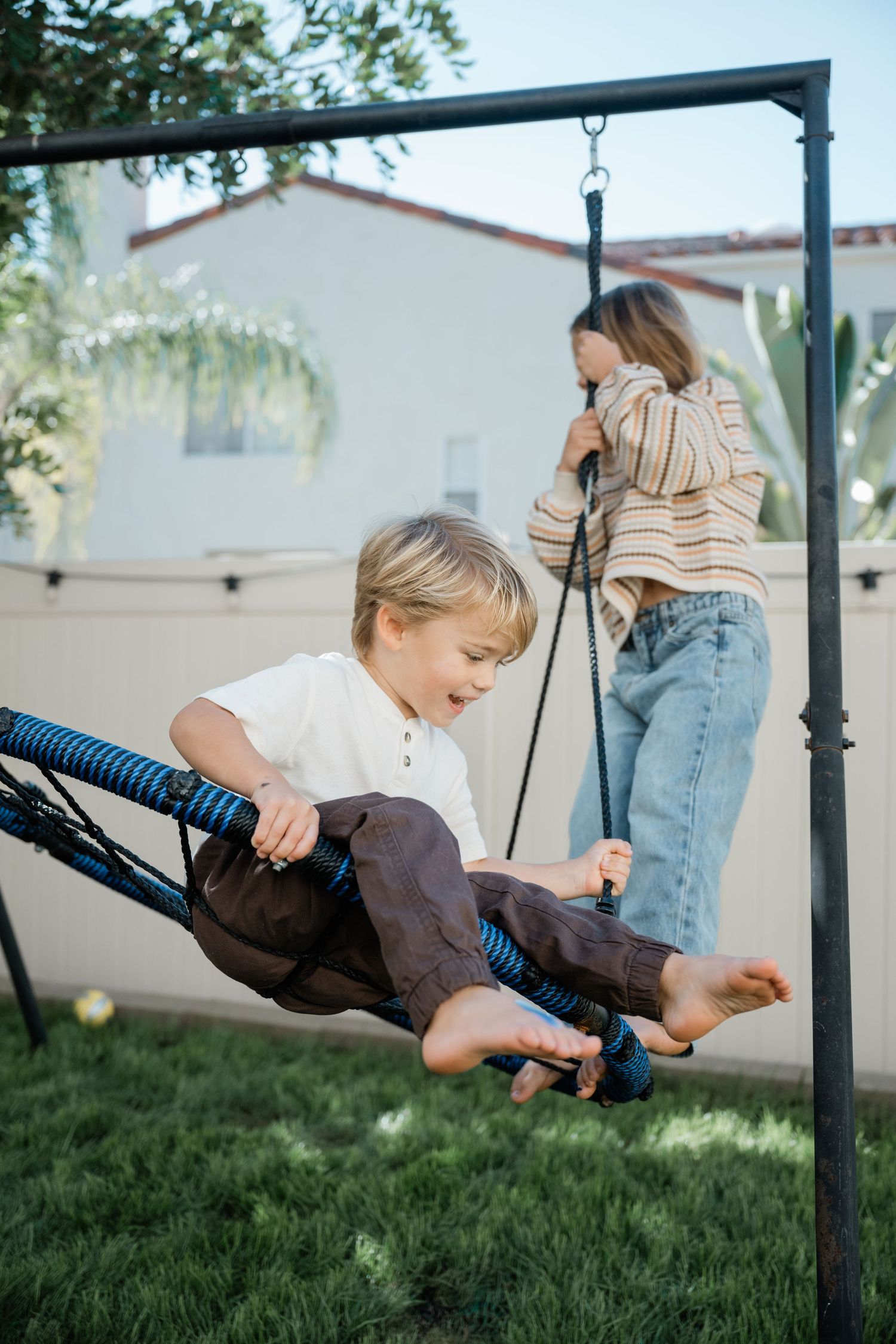 Two children playing on a backyard swing set, with a boy laughing on a spider swing and a girl standing nearby.